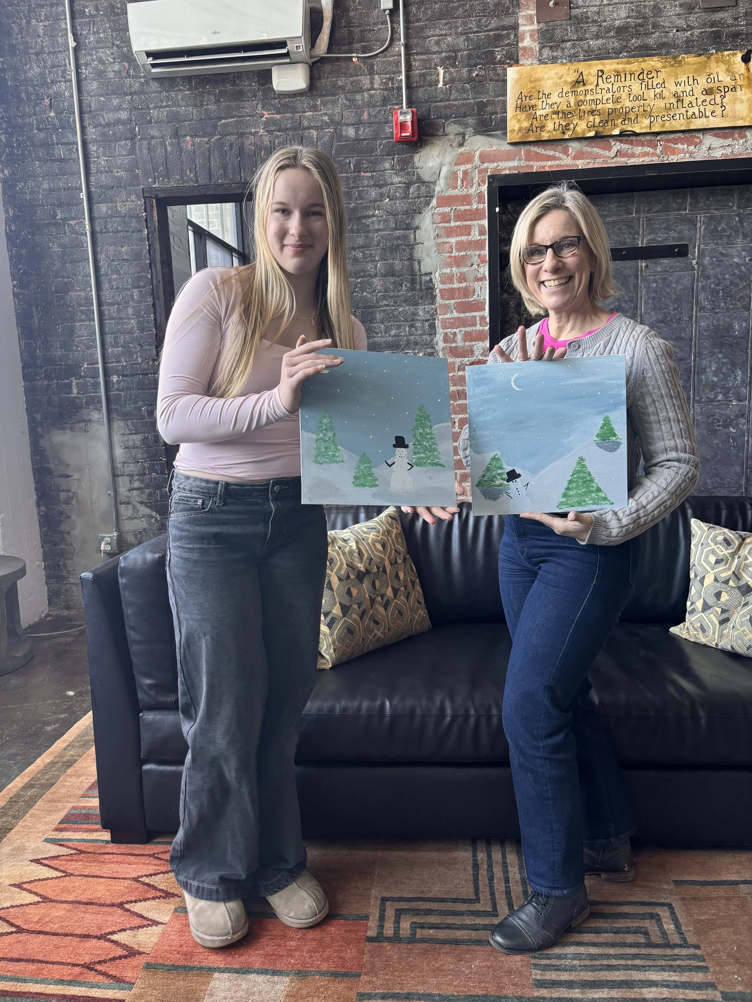 Two women standing indoors holding winter-themed paintings of snowmen and pine trees, smiling at the camera.