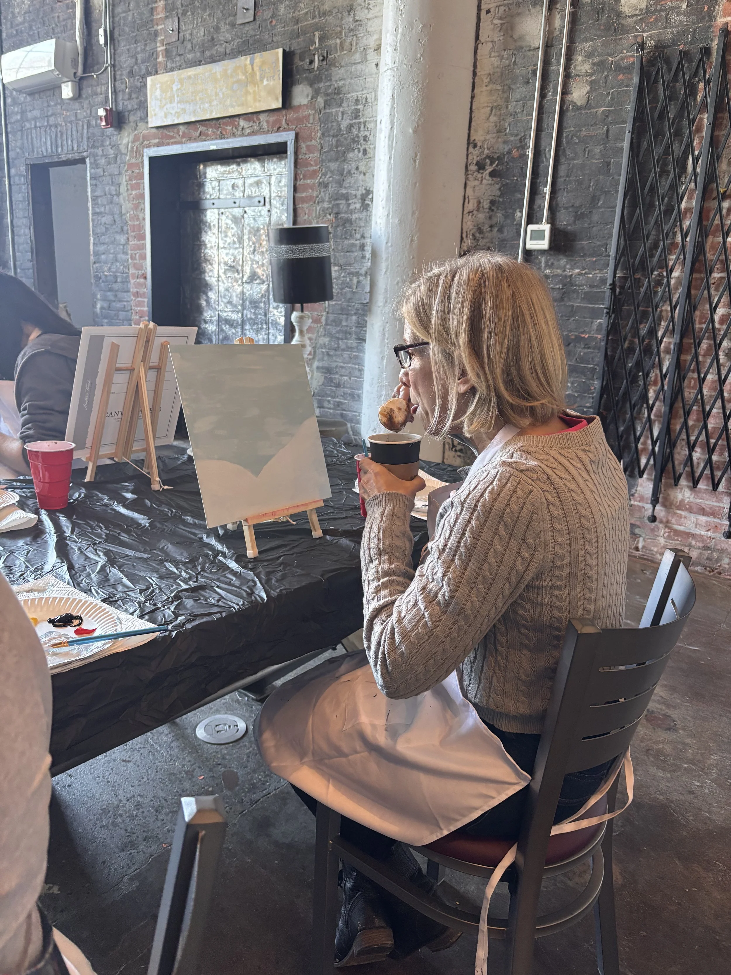 A woman with glasses and blonde hair, wearing a beige sweater and a white apron, is sitting at a table in an industrial-style room with exposed brick walls. She is holding a cup in one hand and a small piece of bread or pastry in her mouth. There are