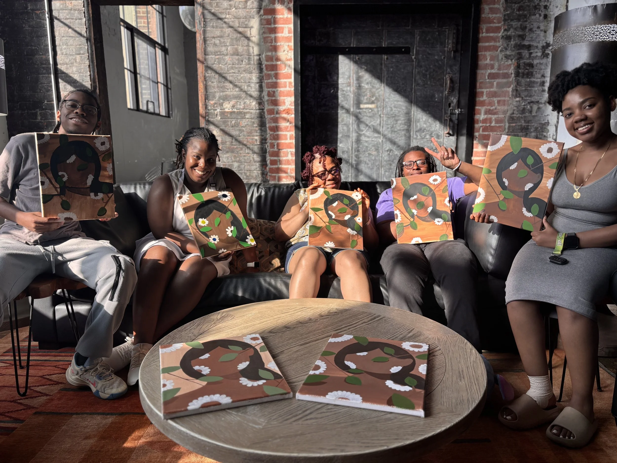 Five women and one man sitting on a black leather sofa, holding colorful paintings with abstract faces and floral patterns, in an industrial-style room with brick walls and large windows. A wooden table in front displays matching paintings.