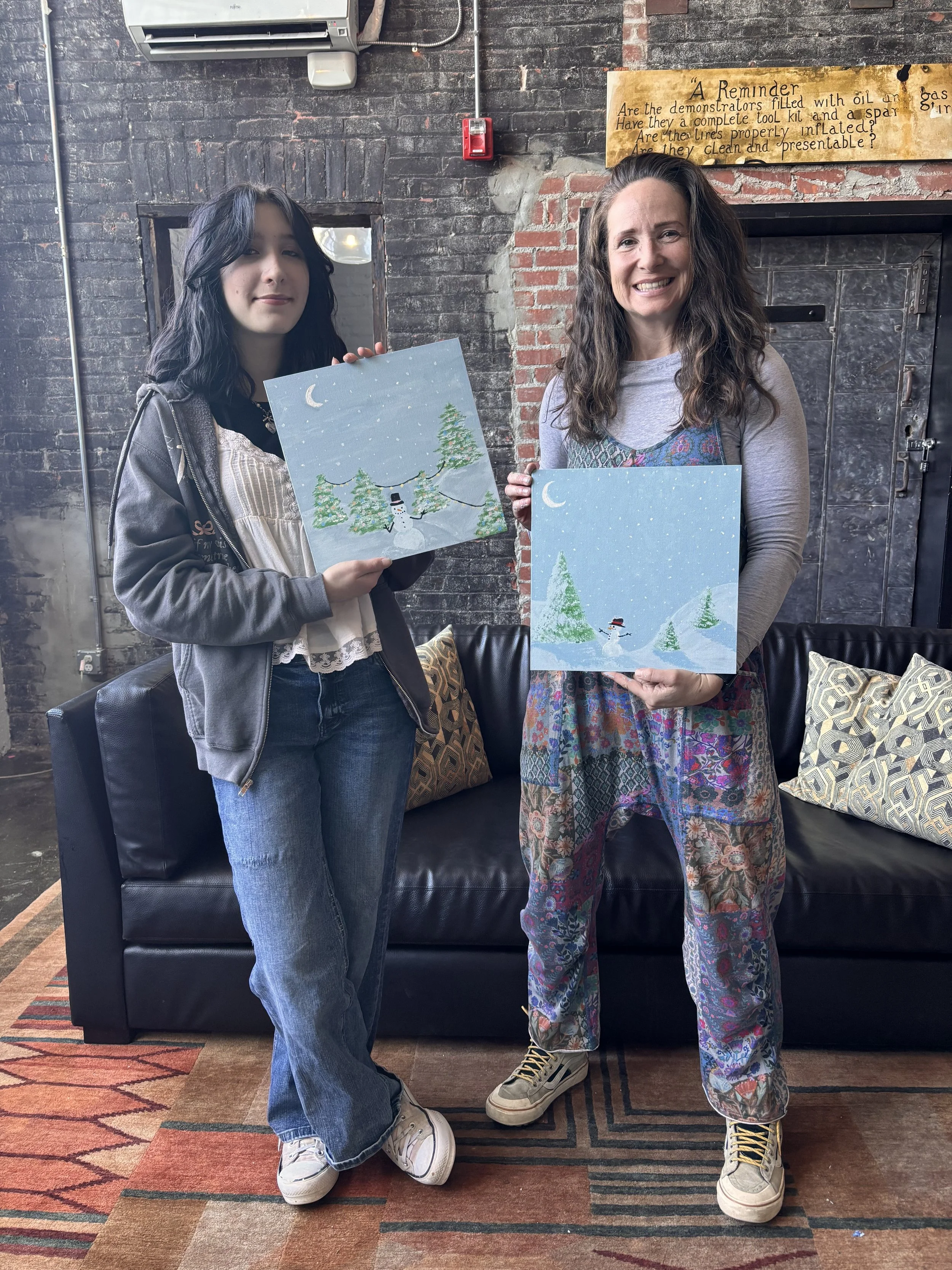 Two women standing indoors, each holding up a winter-themed painting with snowmen, pine trees, and a starry sky. The woman on the left has dark, wavy hair and is wearing a gray hoodie, white blouse, and jeans. The woman on the right has long, curly h