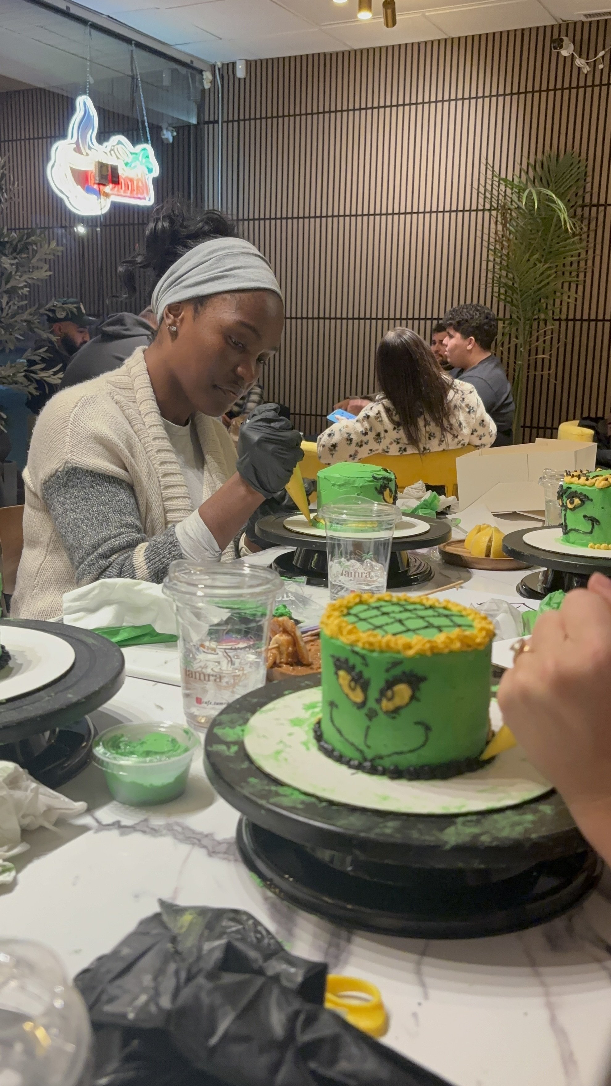 A woman decorating a green cake with yellow and black accents in a restaurant or cafe. Several other decorated cakes are on the table, with people sitting in the background.