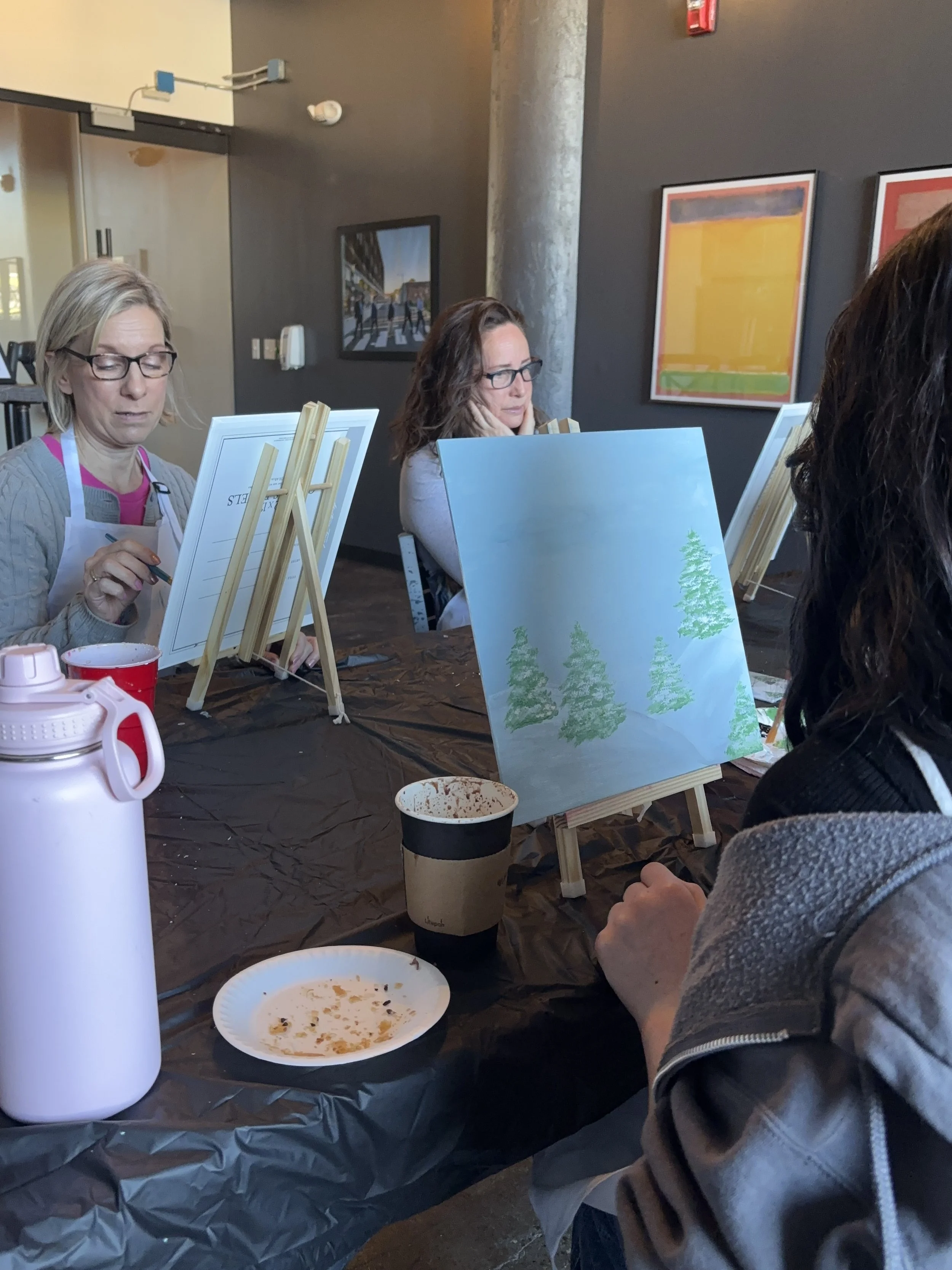 People painting nature scenes on canvases during a group art class at a studio.