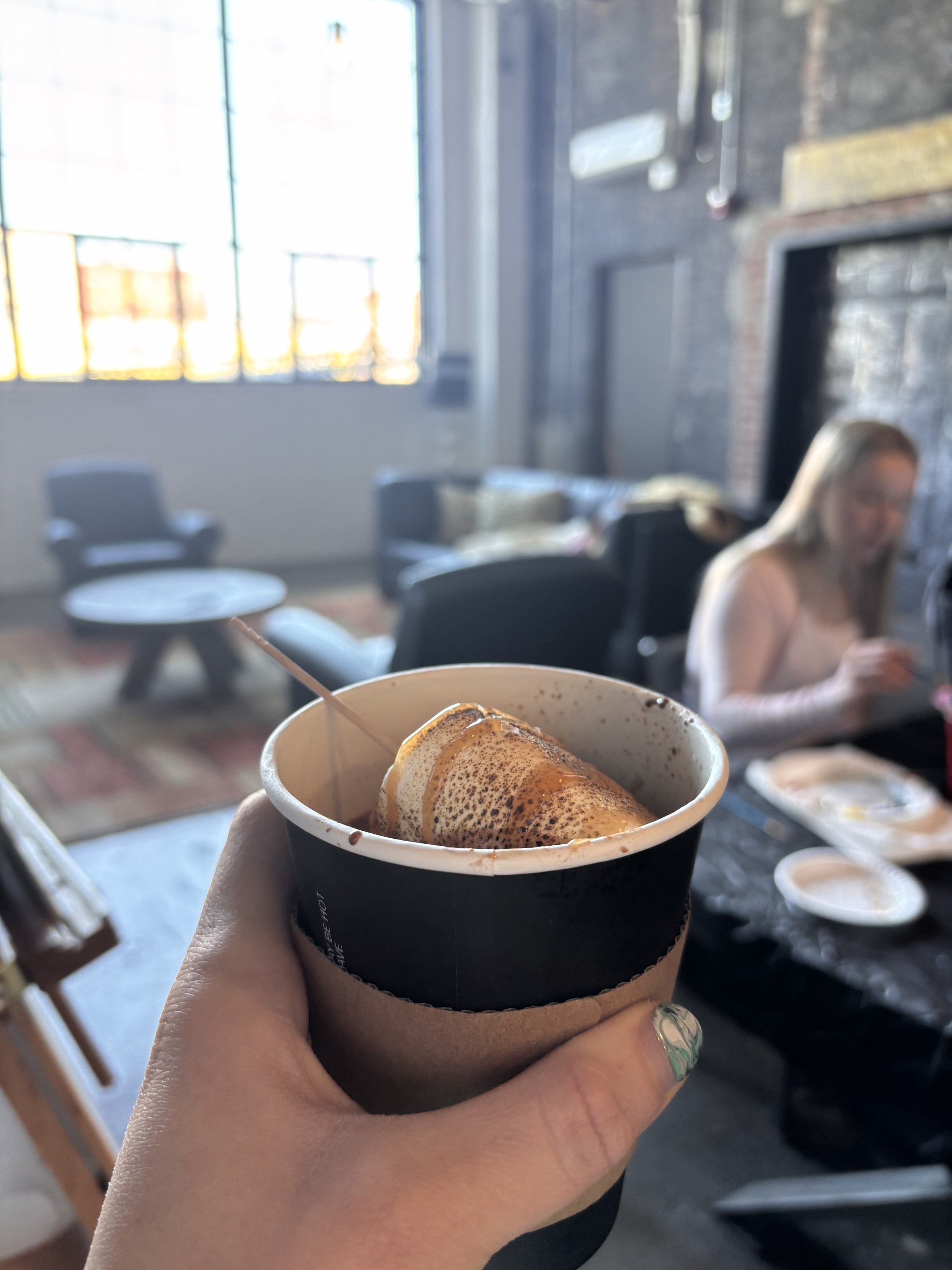 A hand holding a cup of coffee with foam and caramel drizzle inside a black paper cup sleeve, in a cozy industrial-style cafe with a brick wall, large windows, and a woman sitting at a table in the background.