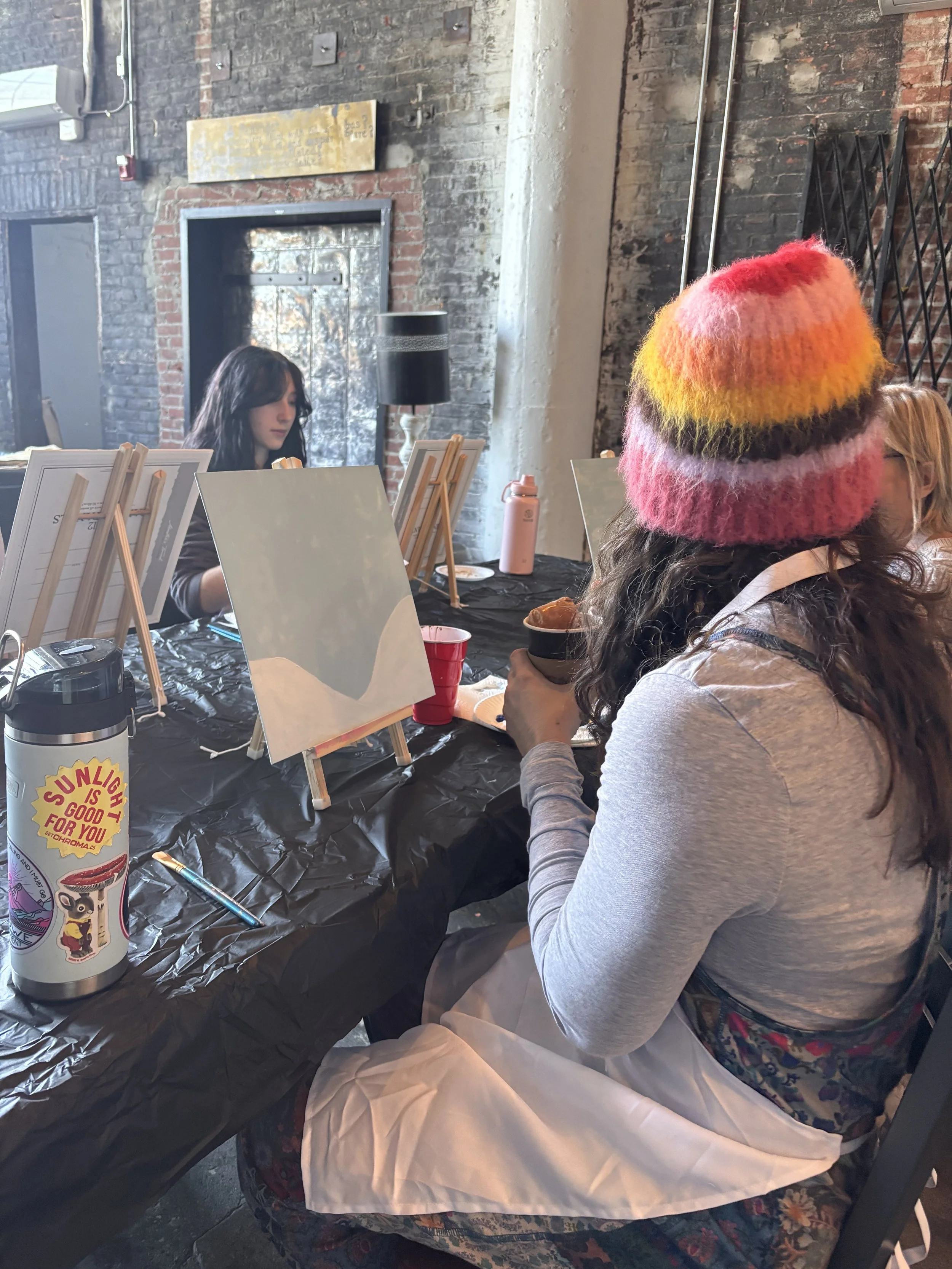 A woman in a colorful knit hat sitting at a table, holding a cup, with other people painting or drawing at an art event in a brick-walled room.