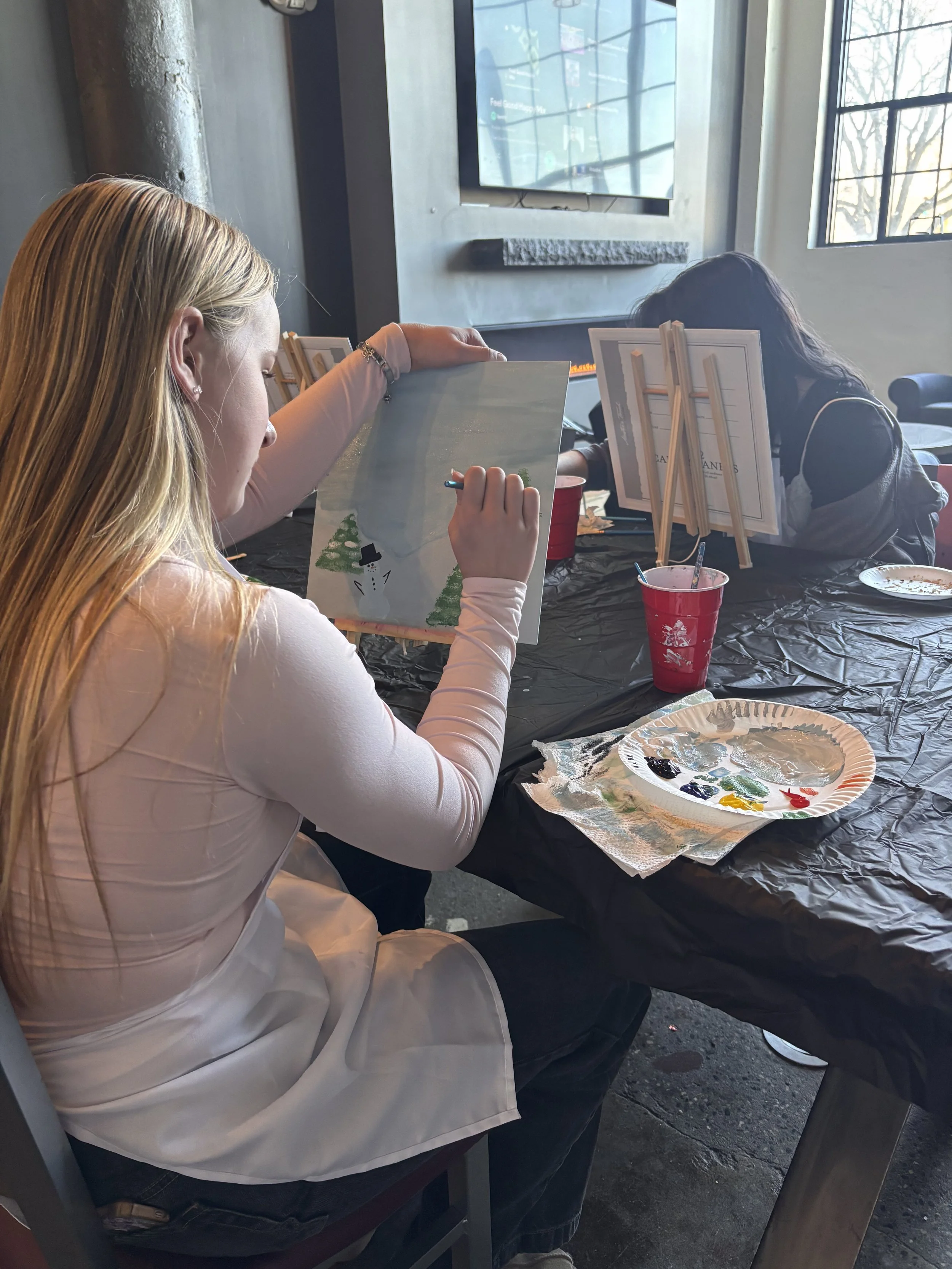 A person painting a winter scene featuring a snowman on a canvas. They are seated at a table covered with a black plastic tablecloth, with a palette of paint and two cups nearby. Two others are also seated at the table, involved in art activity, in a