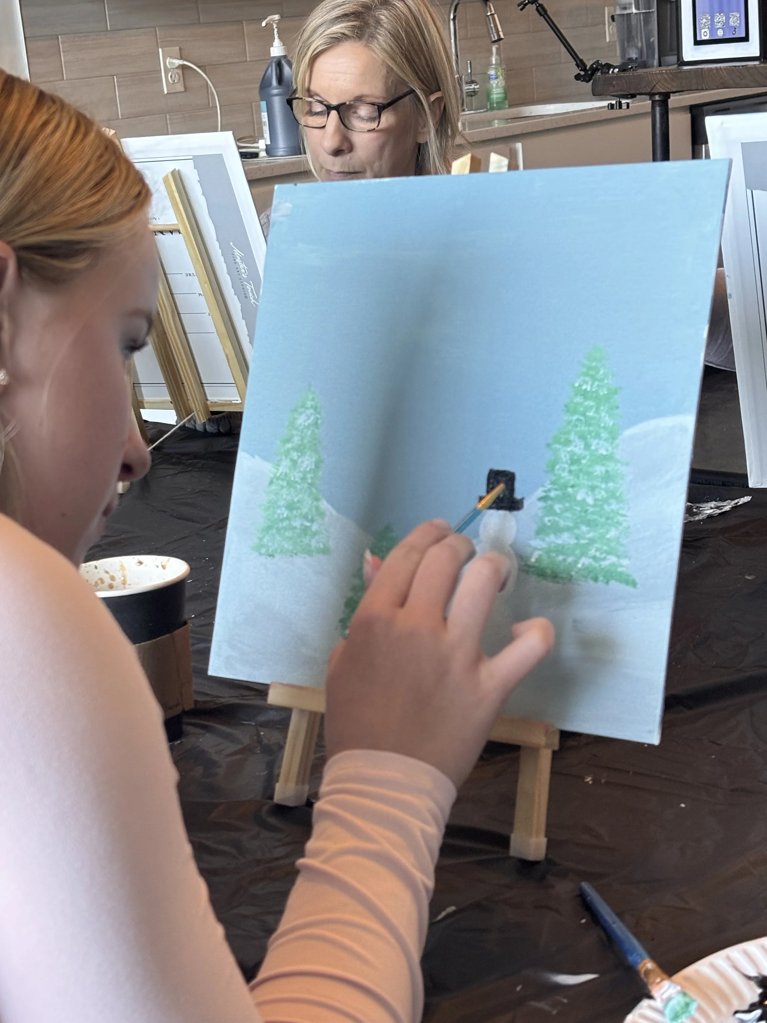 A girl painting a winter scene with snow and two pine trees on a canvas at an art class, with an instructor observing in the background.