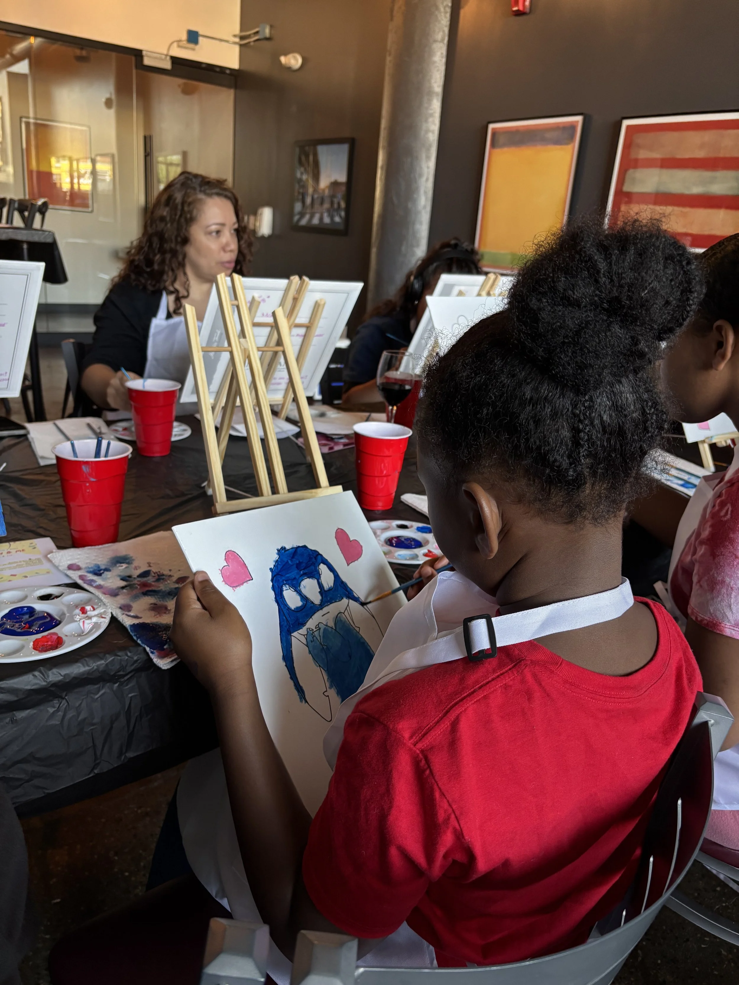A young girl painting a picture of a blue owl with two pink hearts on a canvas, sitting at a table during an indoor art event.