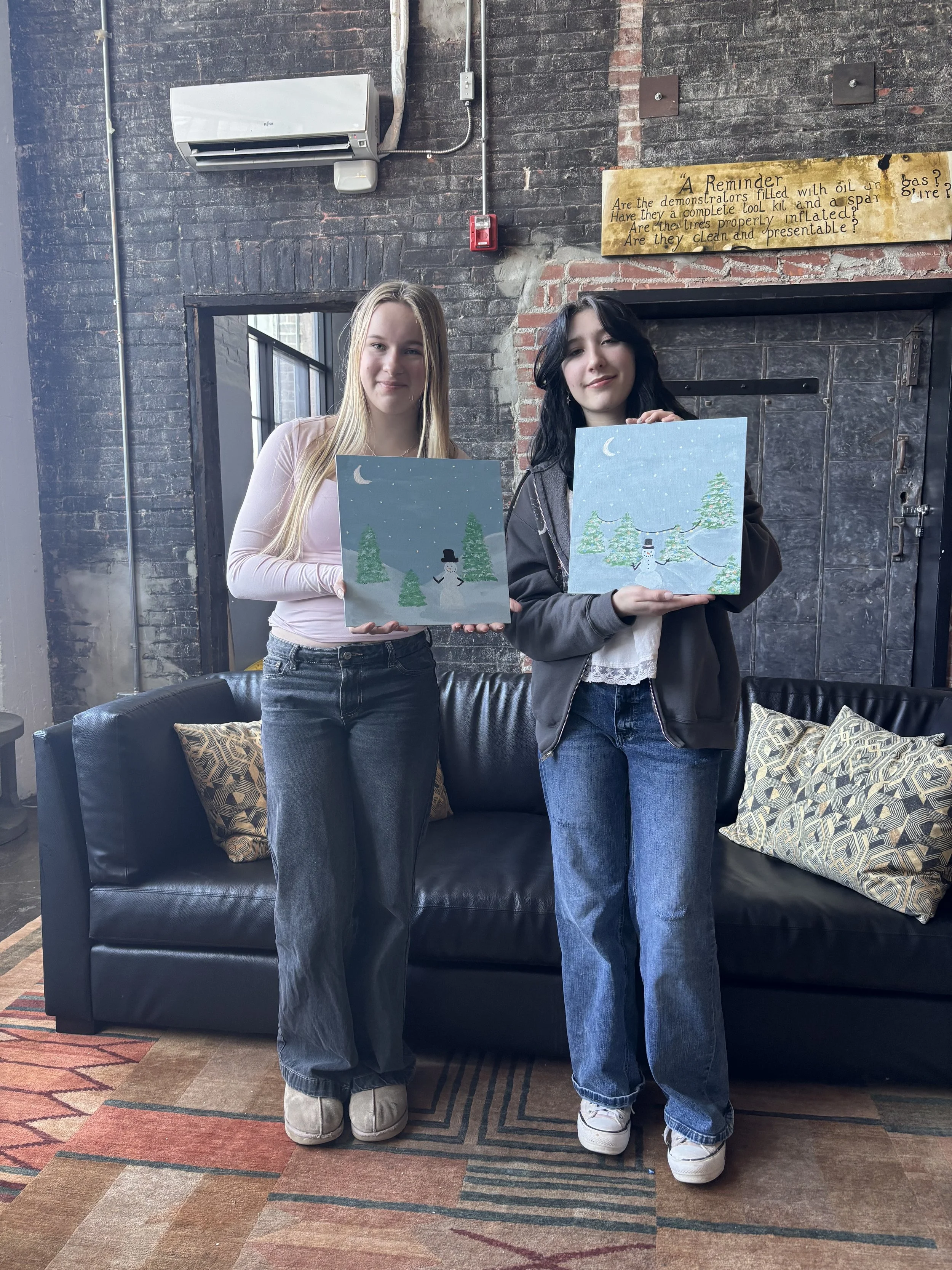 Two young women standing in a room with brick walls, holding paintings of winter scenes with snow, trees, and a snowman, smiling for the camera.