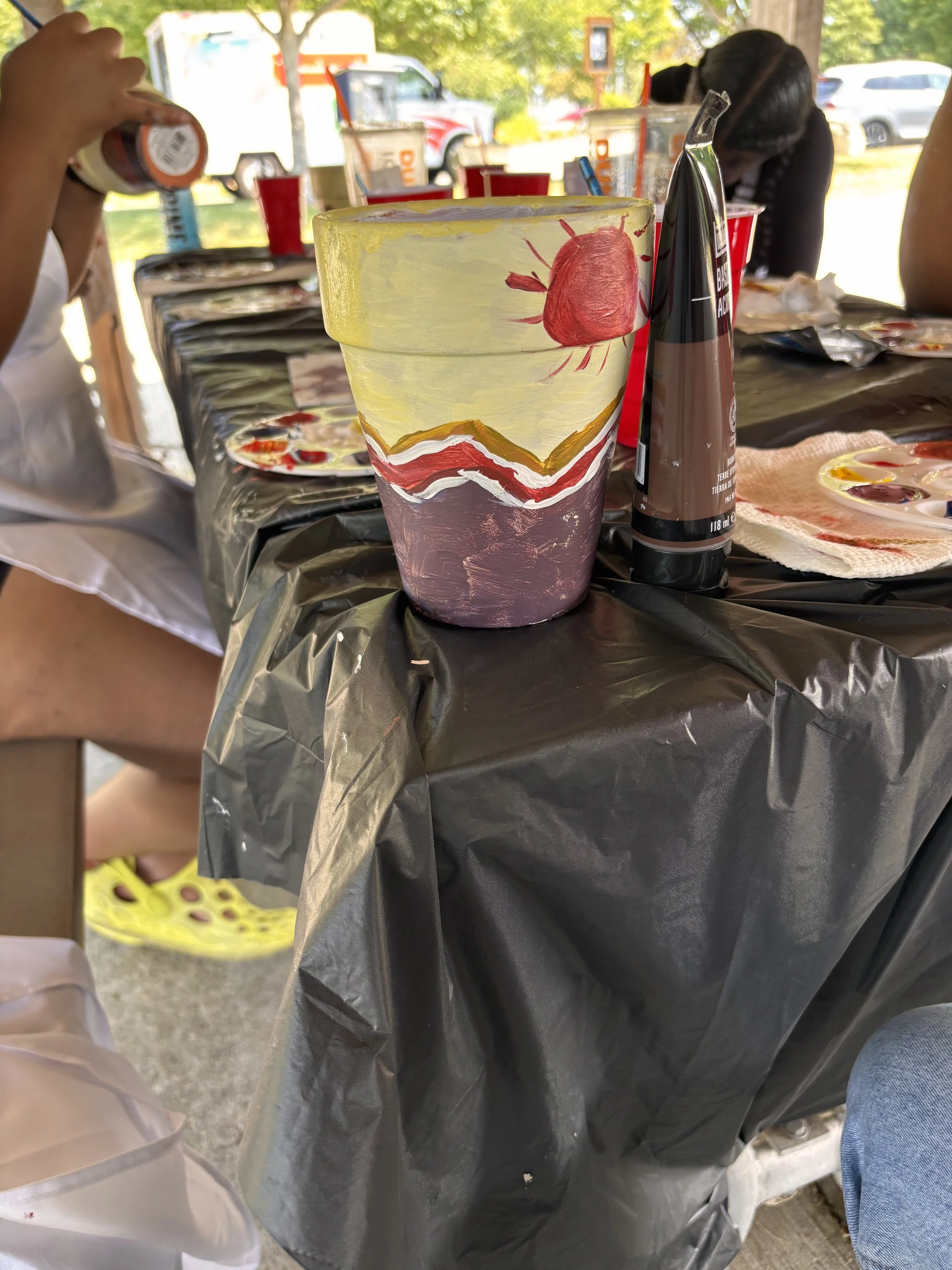 Hand-painted decorated cup with a red sun, yellow sky, yellow and white mountain ranges, and purple ground, placed on a black tablecloth at an outdoor gathering or party.