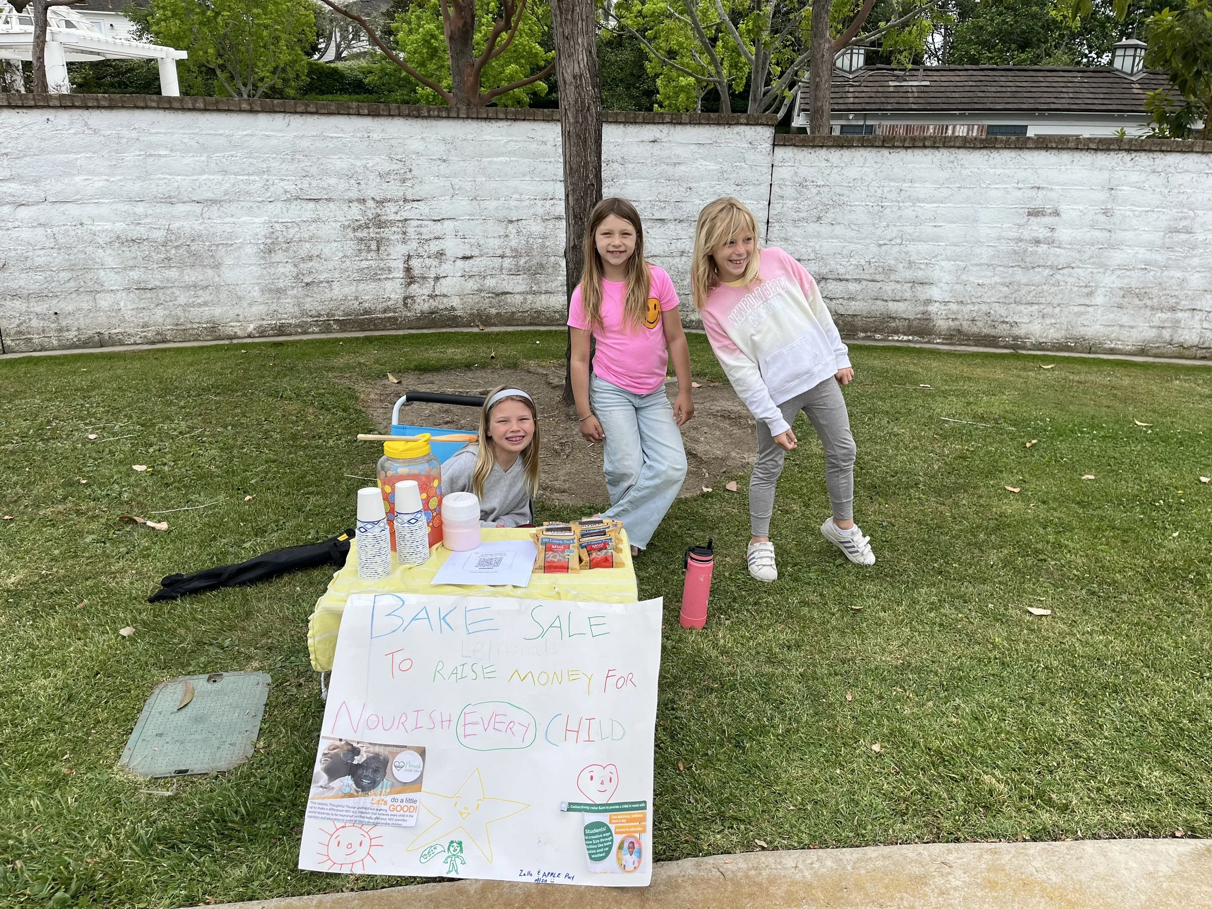 Three young girls at a bake sale table on a grassy yard with a white brick wall behind them. The table has a yellow tablecloth and various baked goods, a jar of candy, and a sign that says "Bake Sale to Raise Money for Nourish Every Child." The girls