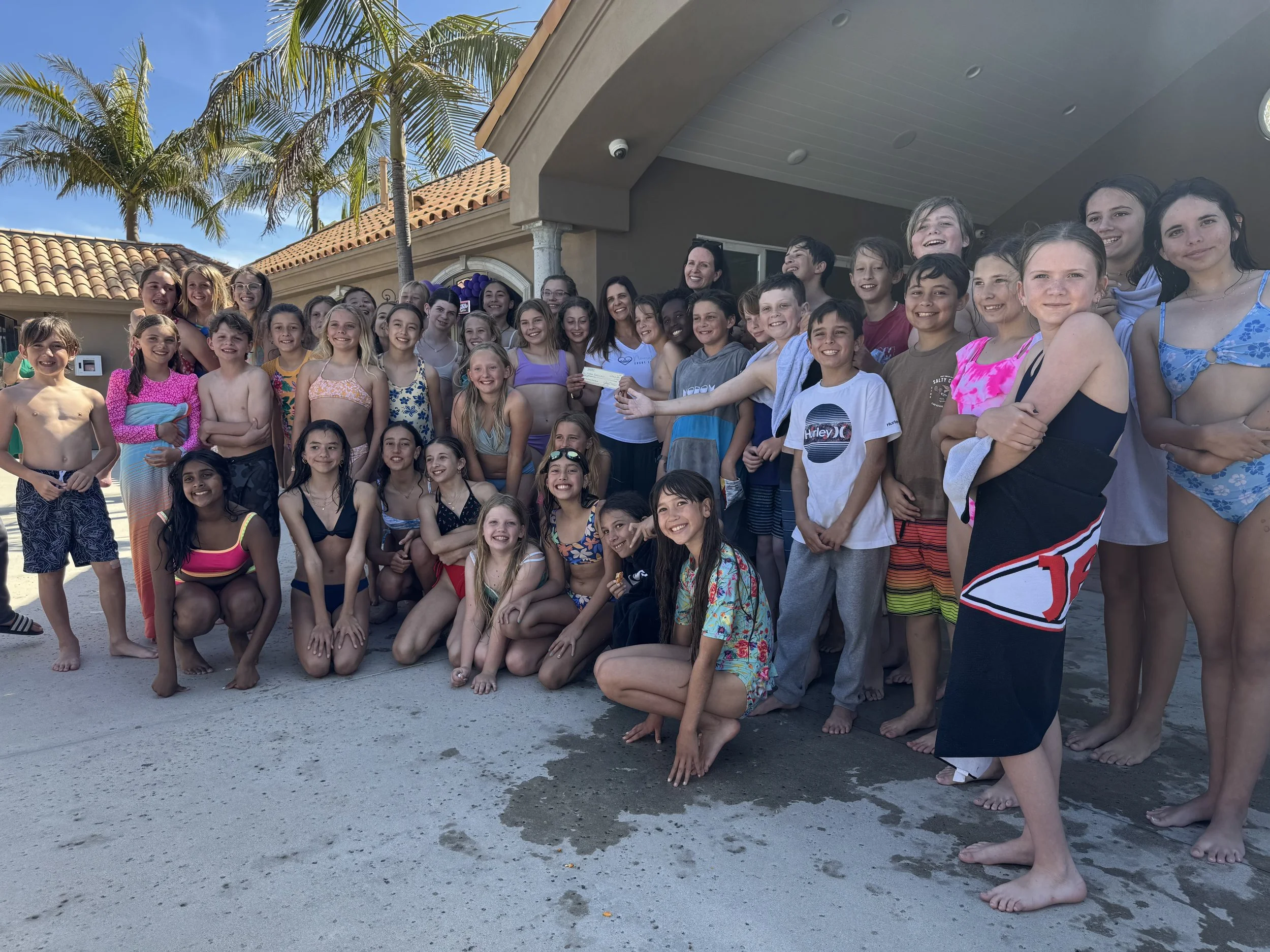 Group of children and teenagers in swimsuits and casual clothing on a sandy area near a pool, smiling and posing for a photo at a sunny outdoor location with palm trees and a building in the background.