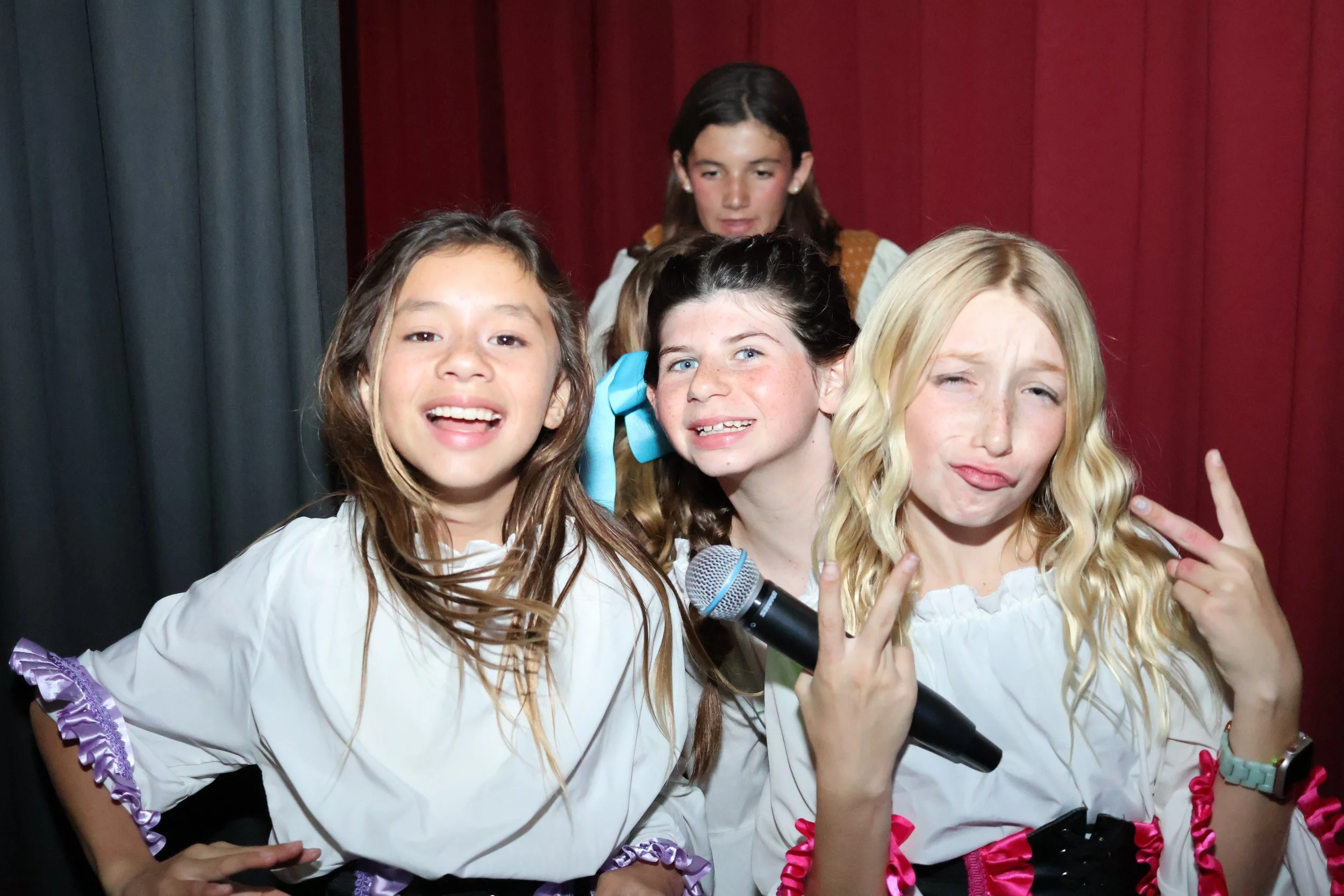 Four young girls on stage, three in the front smiling and making peace signs, one in the back holding a microphone, with a red curtain backdrop.
