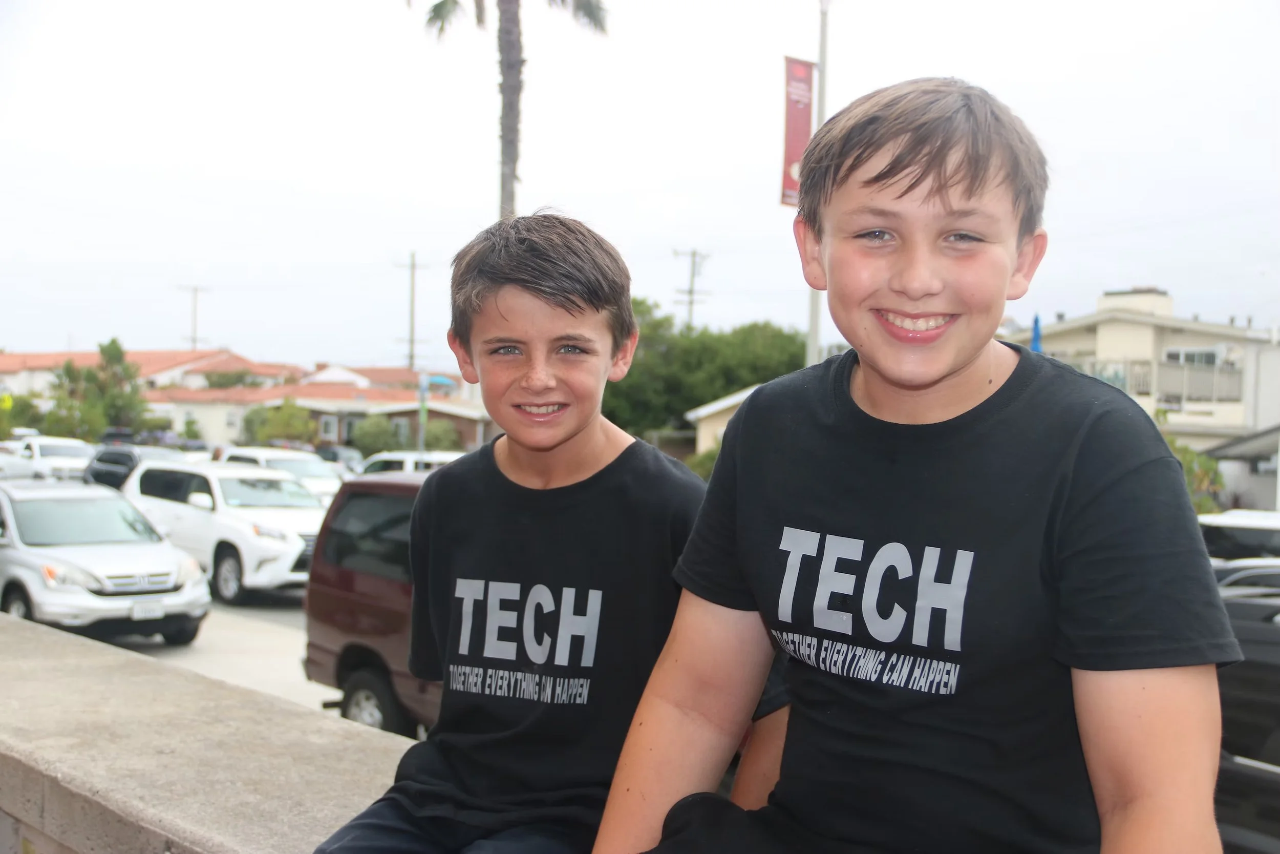 Two young boys sitting outdoors near a parking lot, both wearing black shirts with the word 'TECH' printed on them, smiling at the camera.