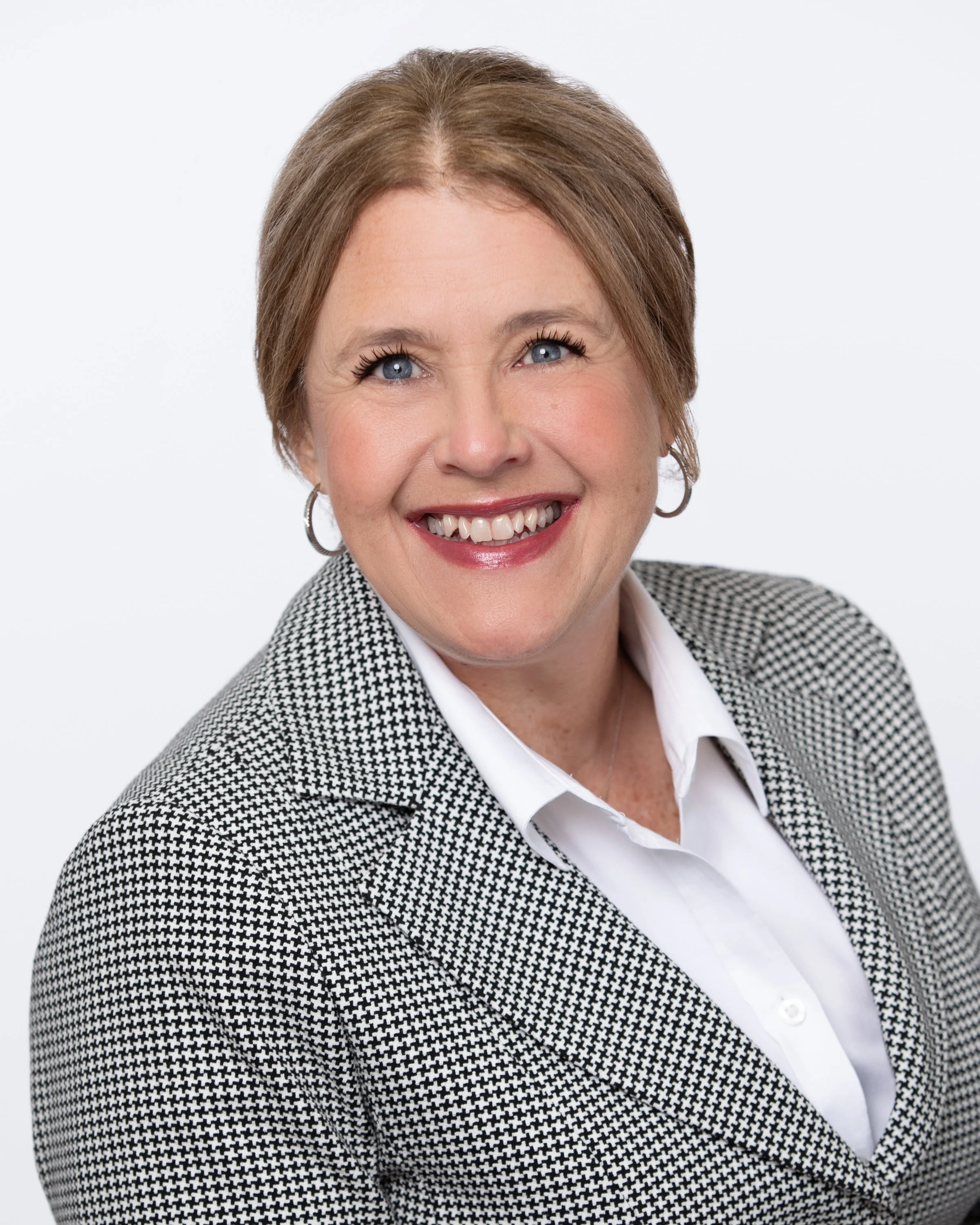 A woman smiling with short light brown hair, blue eyes, wearing a houndstooth blazer over a white shirt, against a plain white background.