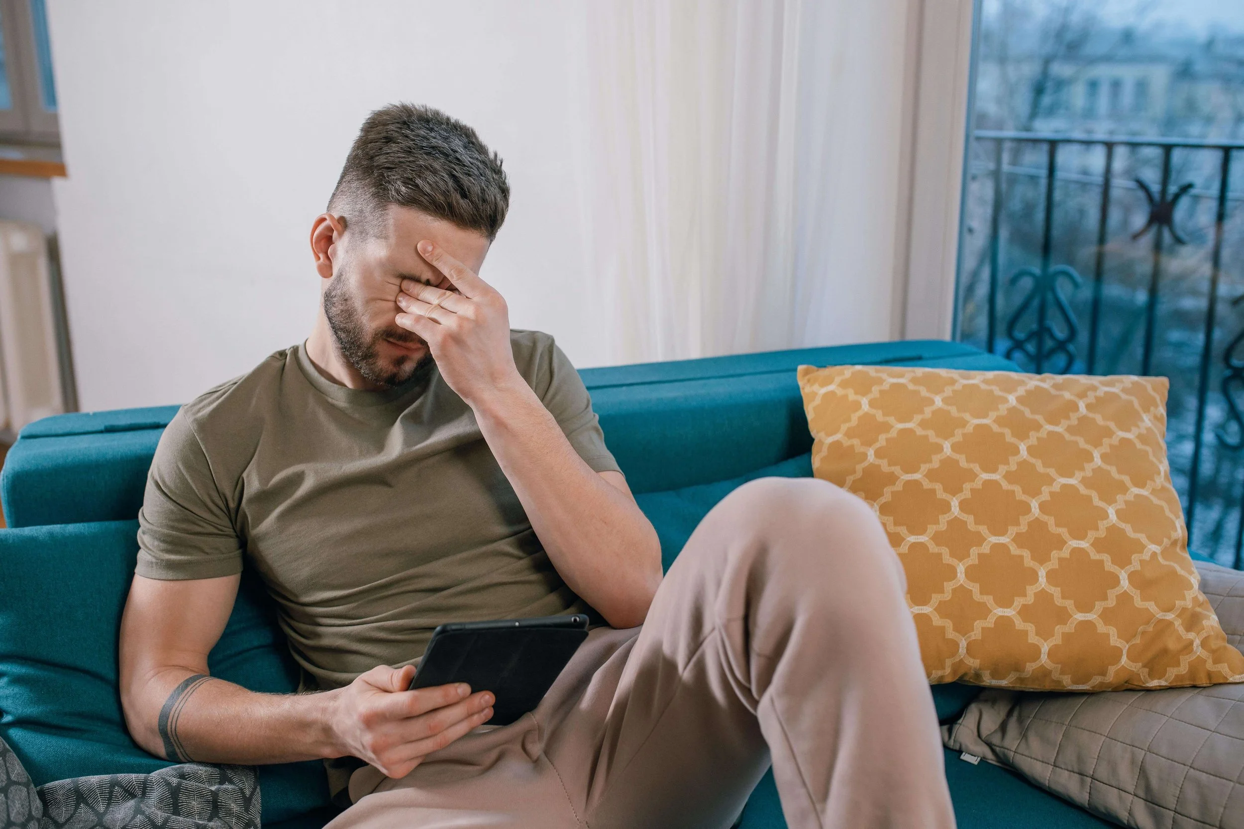 A man sitting on a blue couch holding an Ipad & rubbing his eyes. ERP for ROCD in Boston, MA can help you with uncertainty, doubt in relationships & overthinking. Learn more from an OCD therapist.