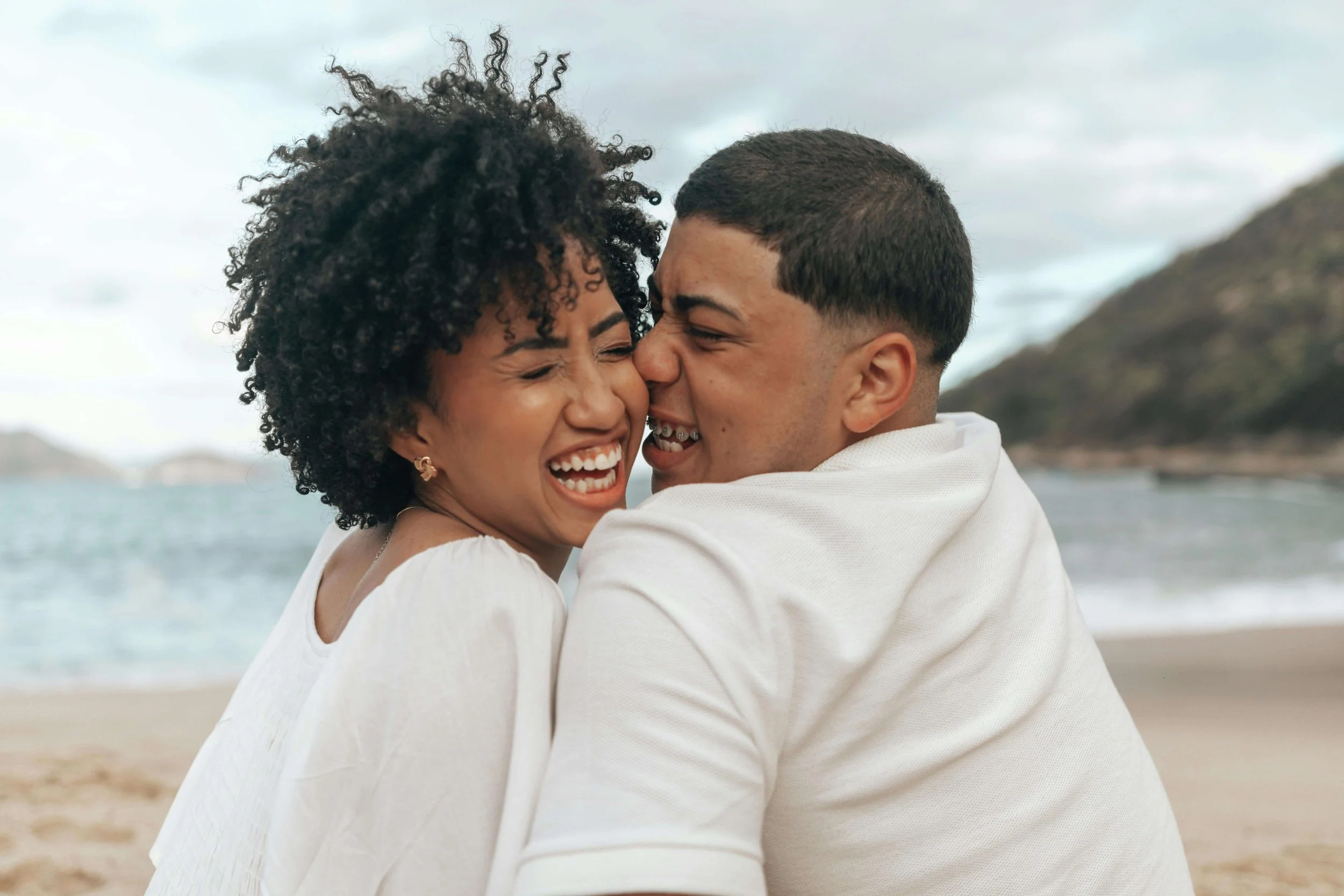 A biracial couple laughing together while siting on the sand near water. An OCd therapist in Boston, MA can help you navigate obsessive thoughts about your relationship, anxiety & more. Reach out today to get started.