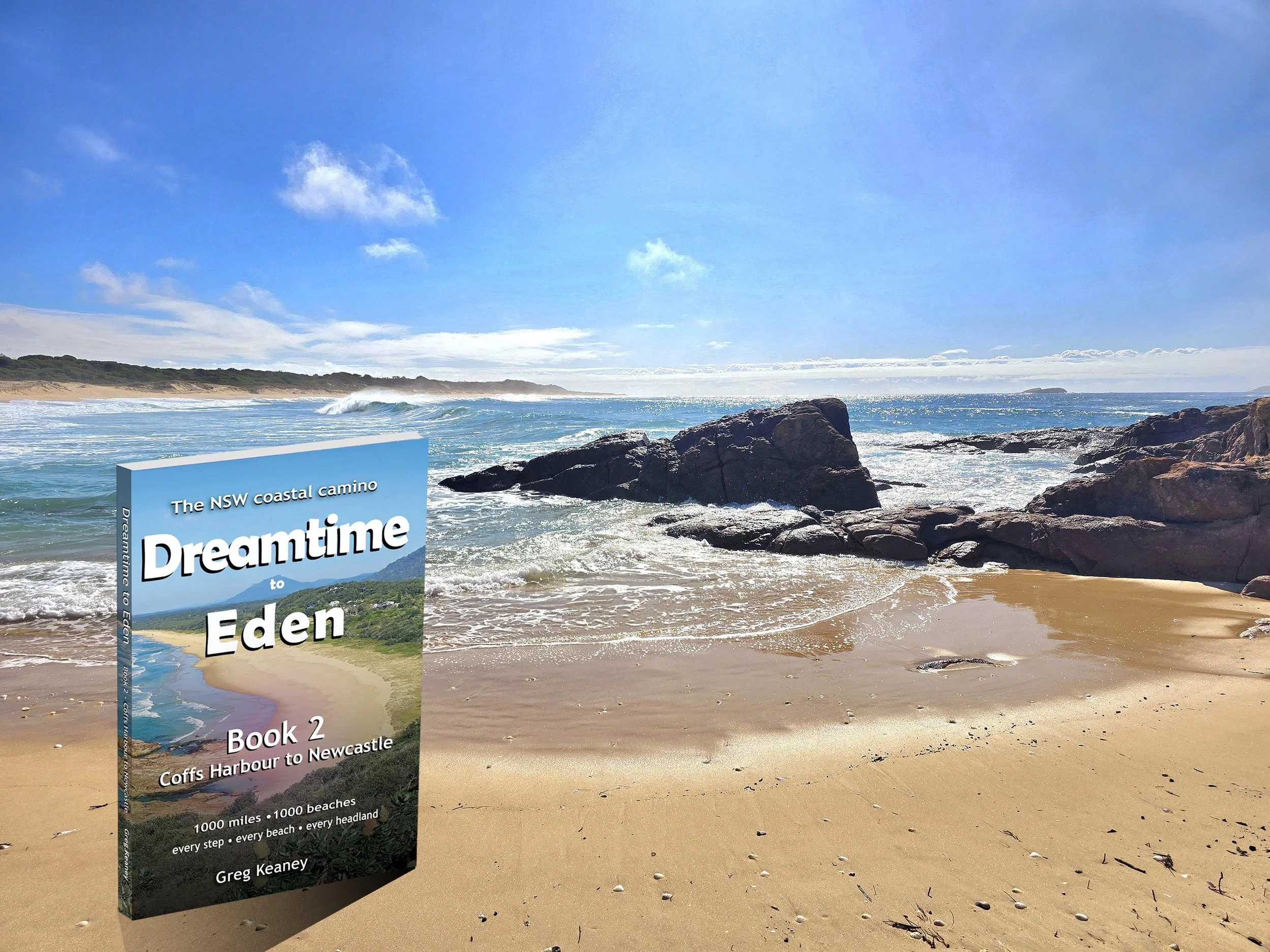 A scenic coastal beach with rocks in the foreground, ocean waves, a sandy shoreline, and a partly cloudy blue sky. There is a book titled 'Dreamtime to Eden, Book 2' by Greg Keaney standing upright on the beach.