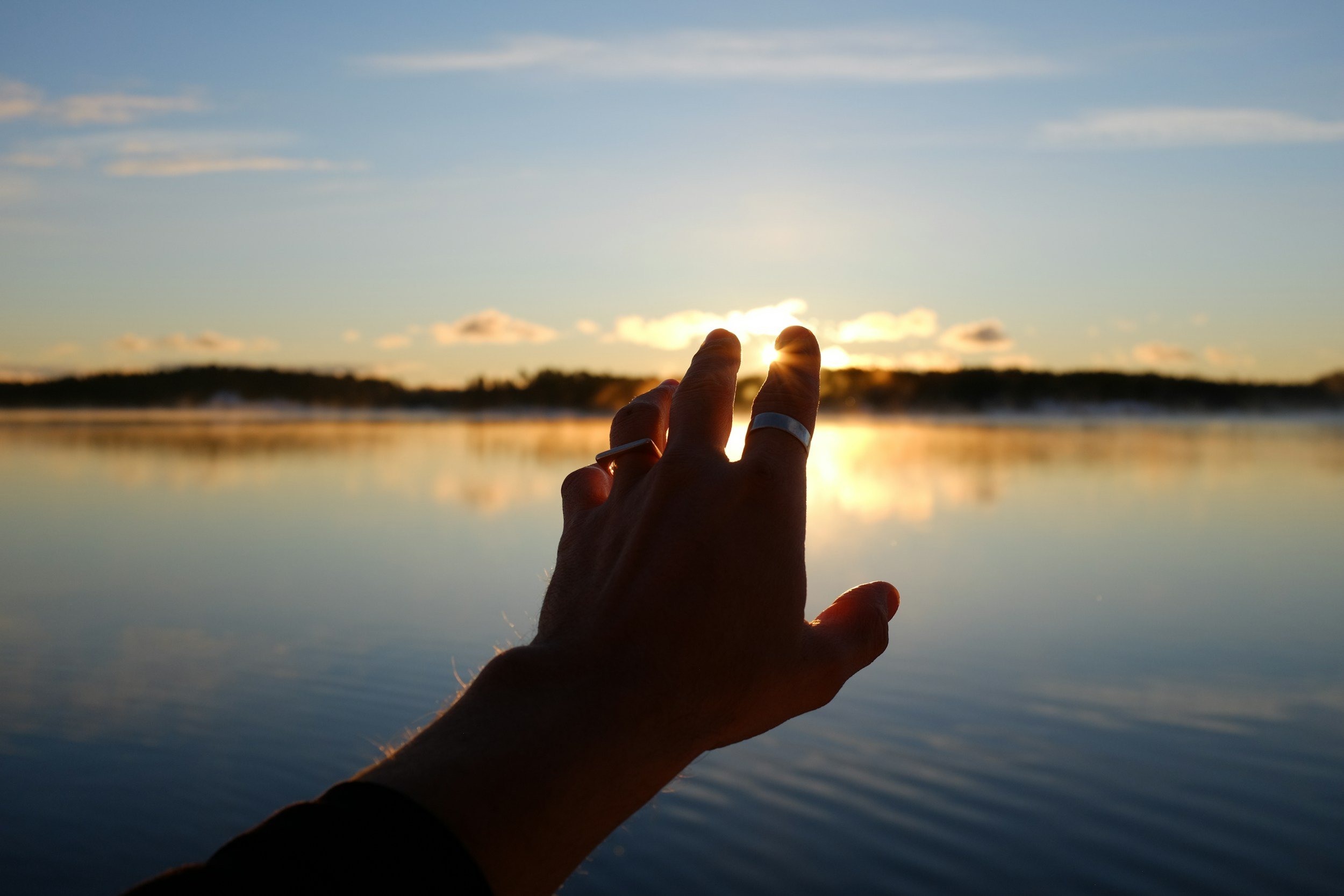 Person's hand reaching towards the sunset over a reflective body of water, with a scenic landscape in the background.