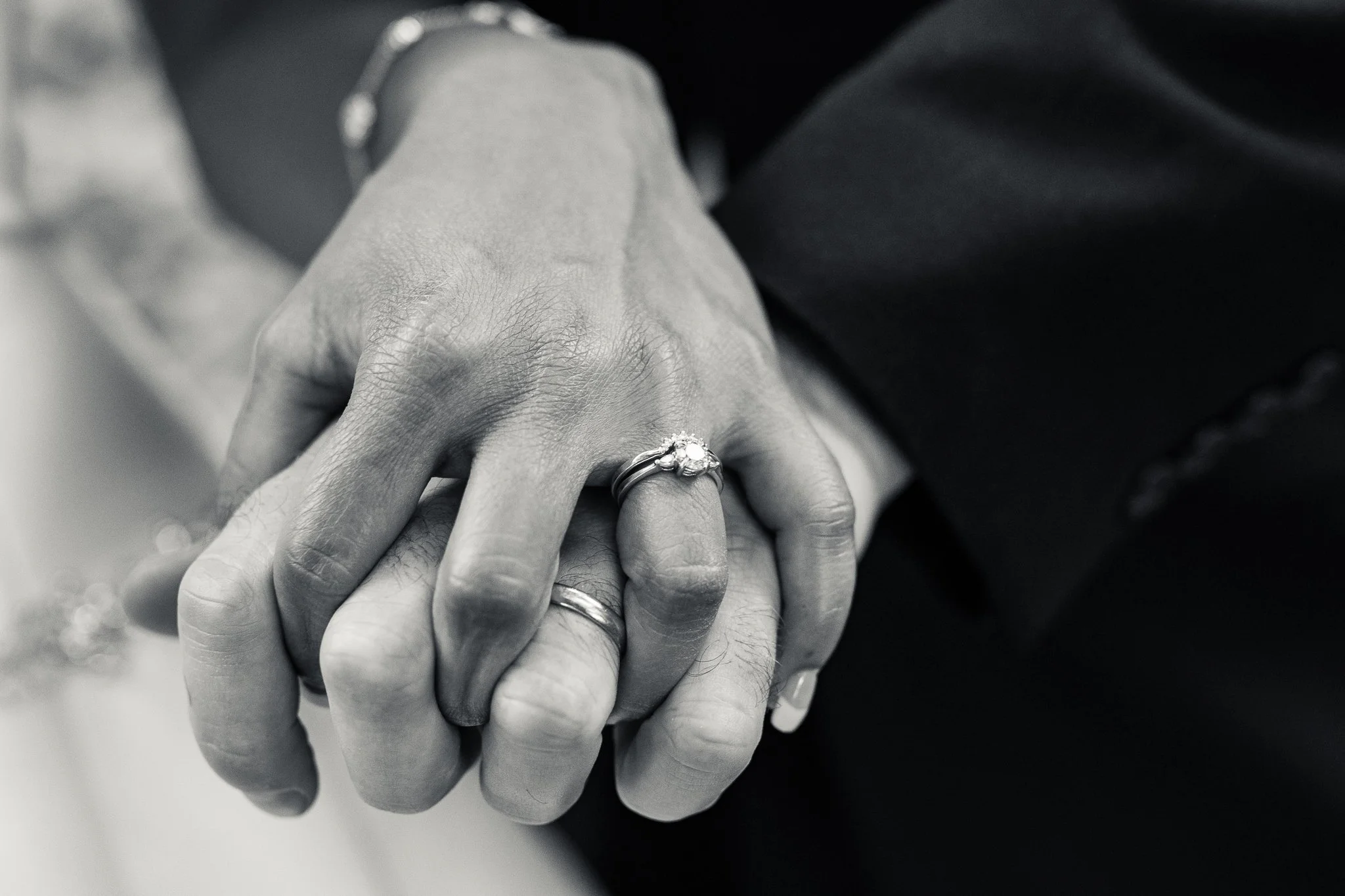 Close-up of two hands holding each other, showing wedding rings, in black and white.
