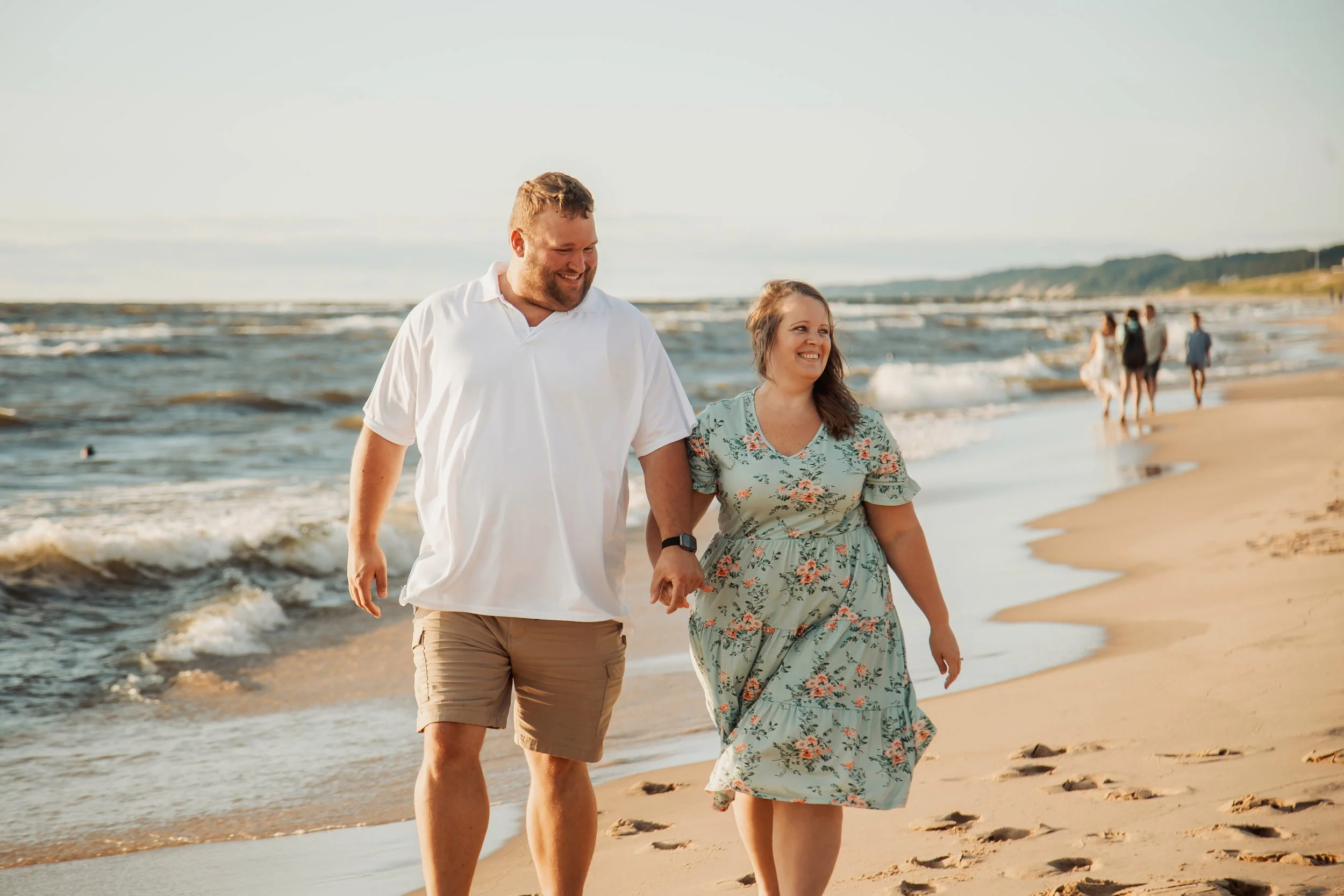 A couple walking hand in hand on the beach, smiling, with ocean waves in the background and other people walking along the shoreline.