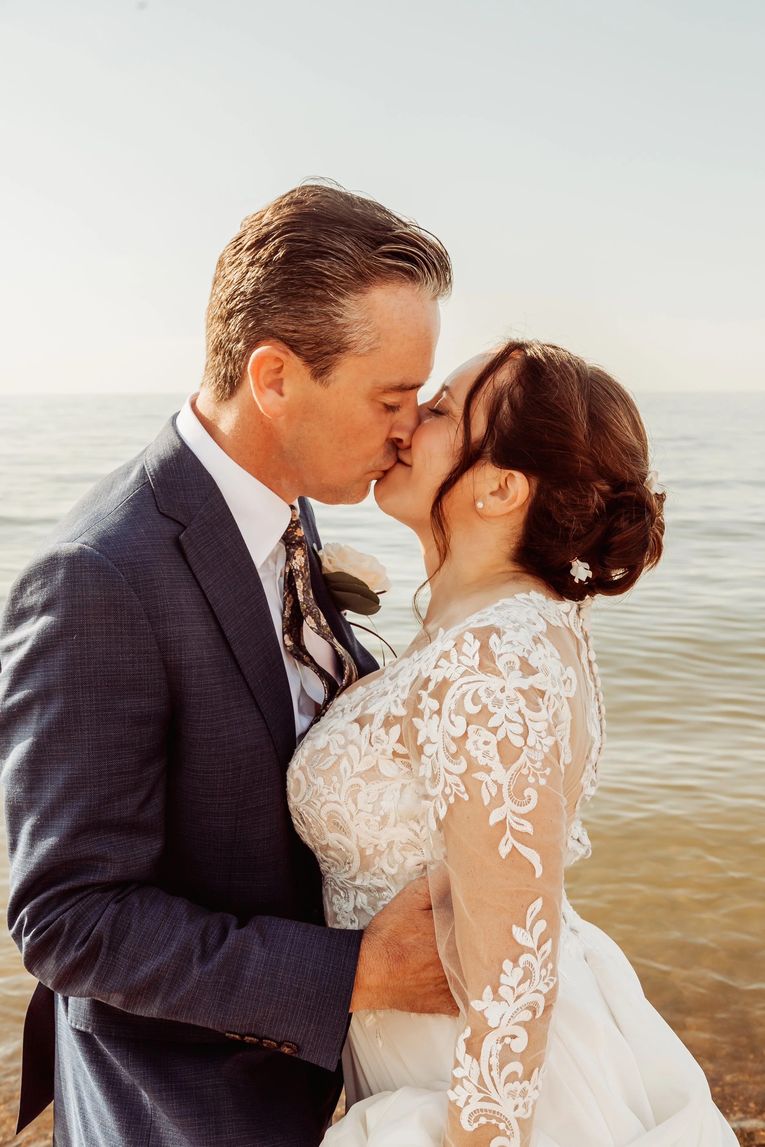 A bride and groom share a kiss on a beach, with water in the background, during their wedding.