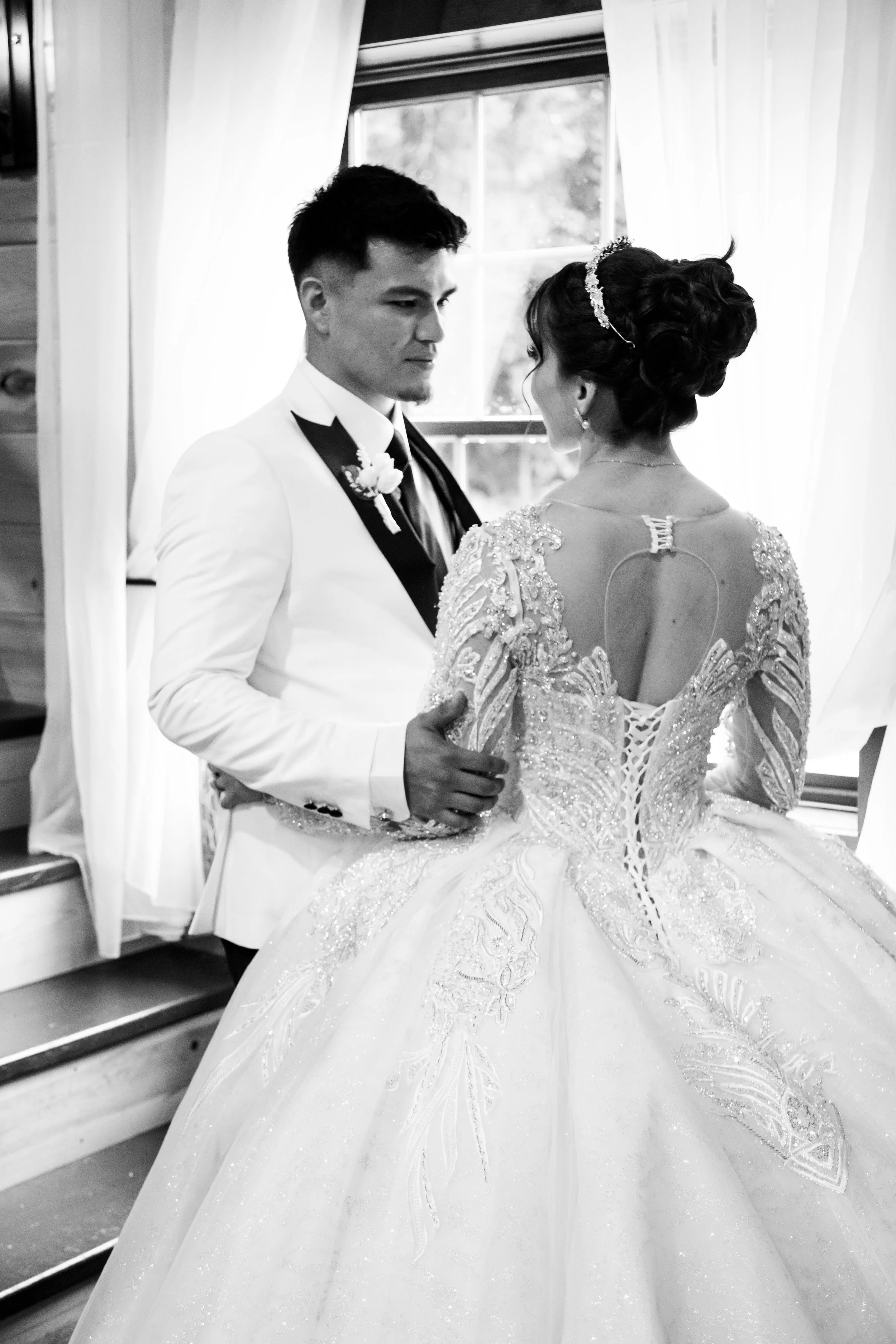 A black and white photo of a bride and groom facing each other indoors near a window with curtains, with the groom holding the bride's waist. The bride wears an ornate wedding gown with intricate embroidery, and the groom wears a tuxedo with a bow ti