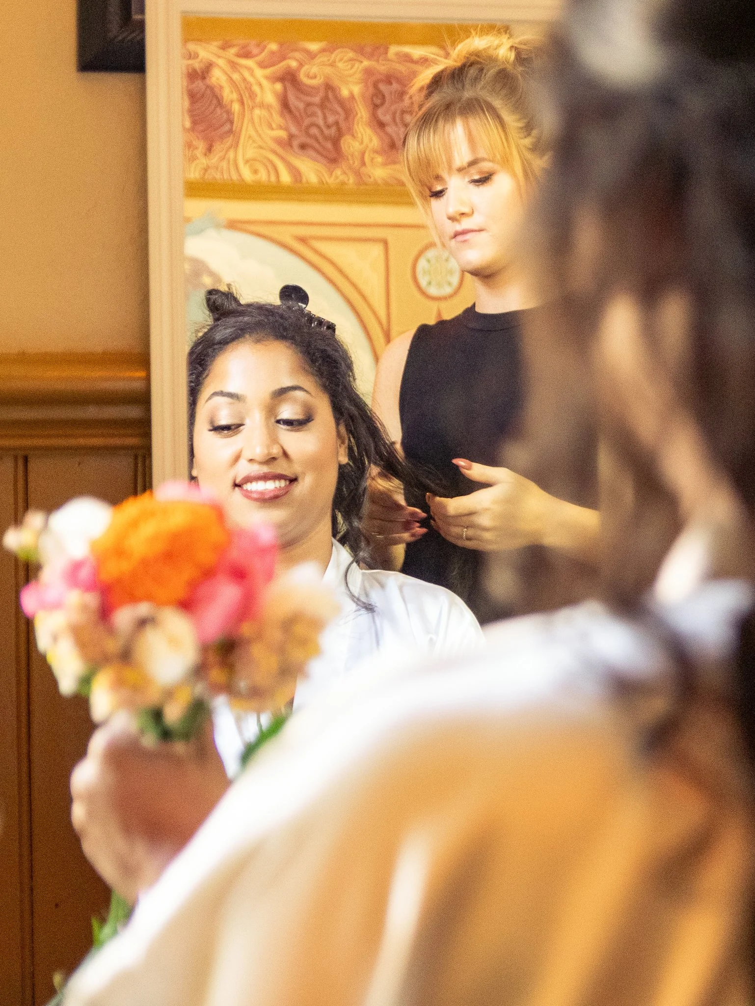 A woman with dark hair getting her hair styled, holding a bouquet of flowers, and smiling. A hairstylist with blonde hair is working nearby, in a room decorated with ornate wallpaper and warm lighting.