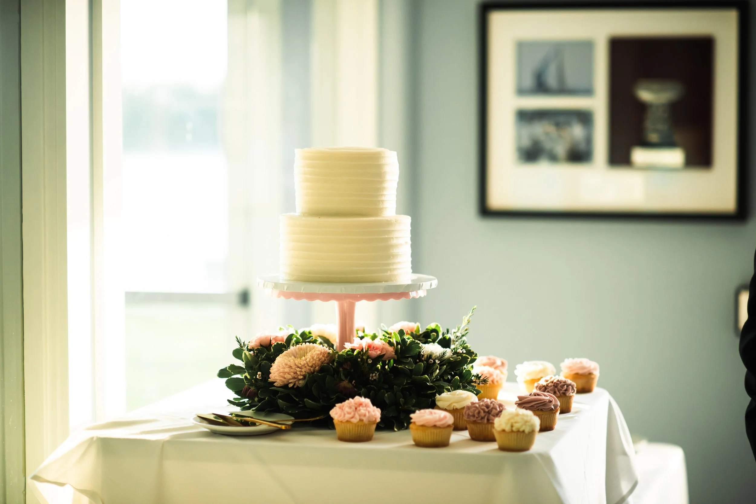 A white, two-tiered frosted cake on a pink stand surrounded by pink and white cupcakes and green foliage on a table with a white tablecloth, near a window with sunlight.
