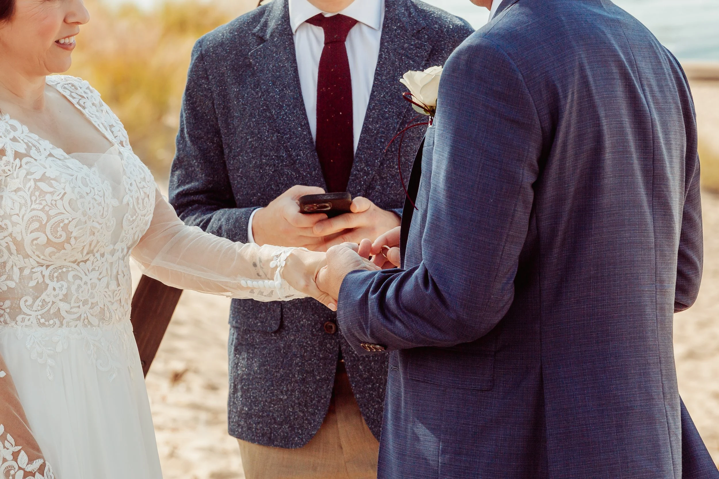 A couple getting married, holding hands and facing each other during the ceremony, with an officiant in the background holding a phone.