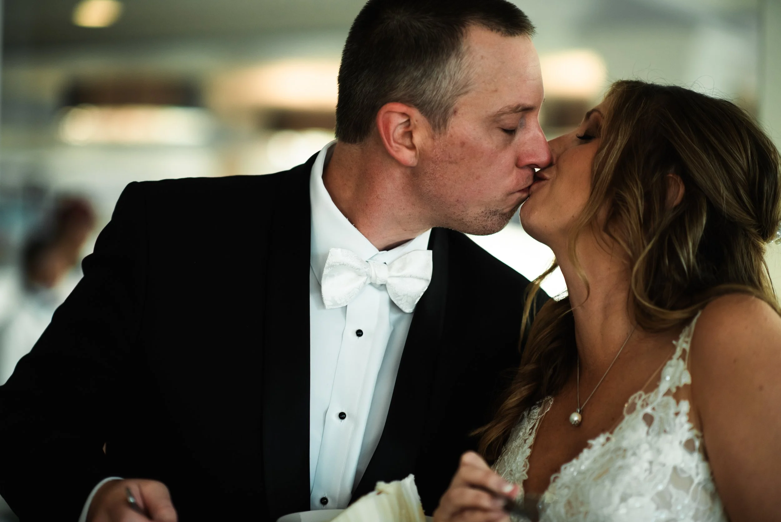 A man and woman are kissing at a wedding reception. The man is wearing a black tuxedo with a white shirt and bow tie, and the woman is in a white lace dress with a pearl necklace.