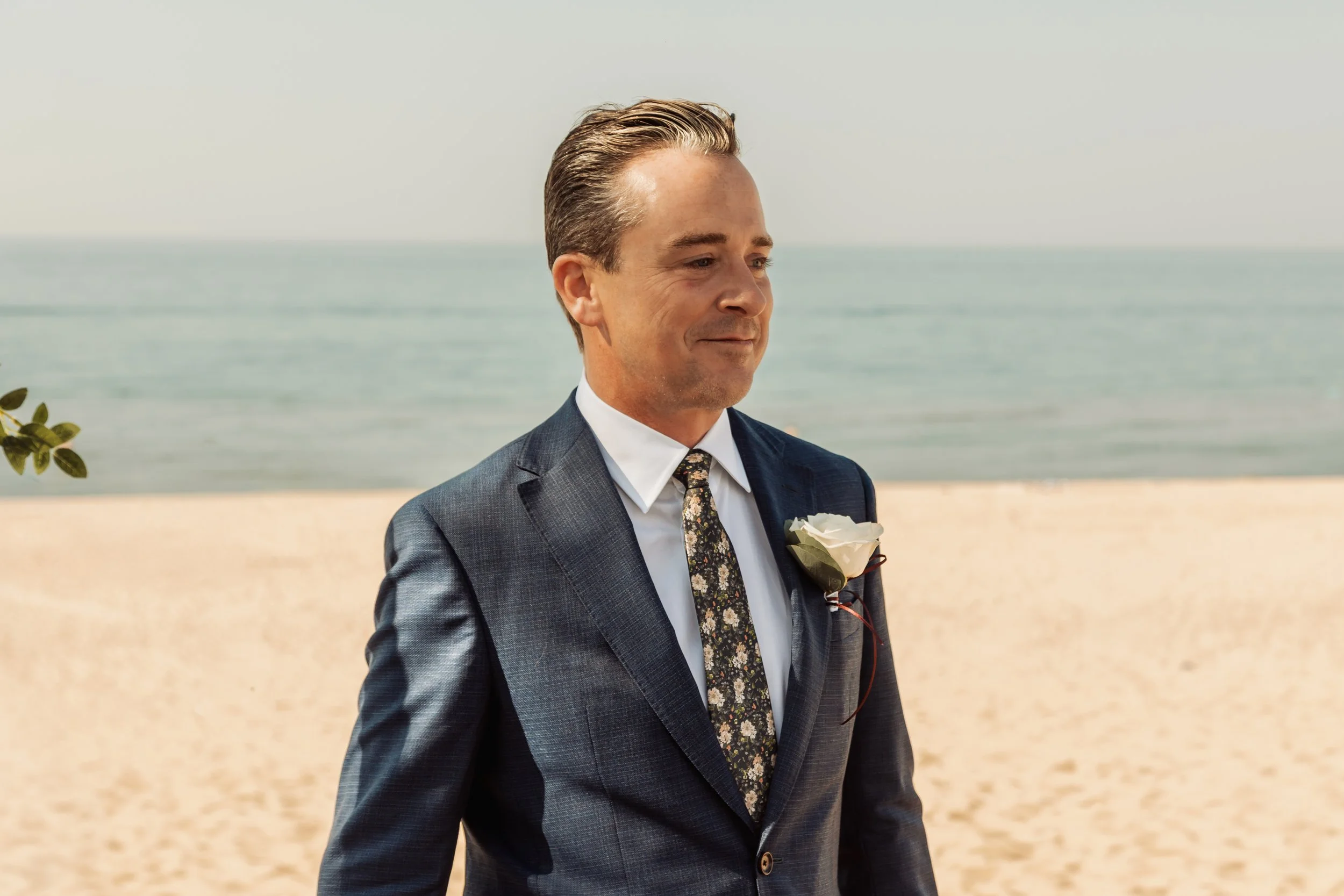 A man in a suit with a floral tie and a white rose boutonniere standing on a sandy beach with the ocean in the background.