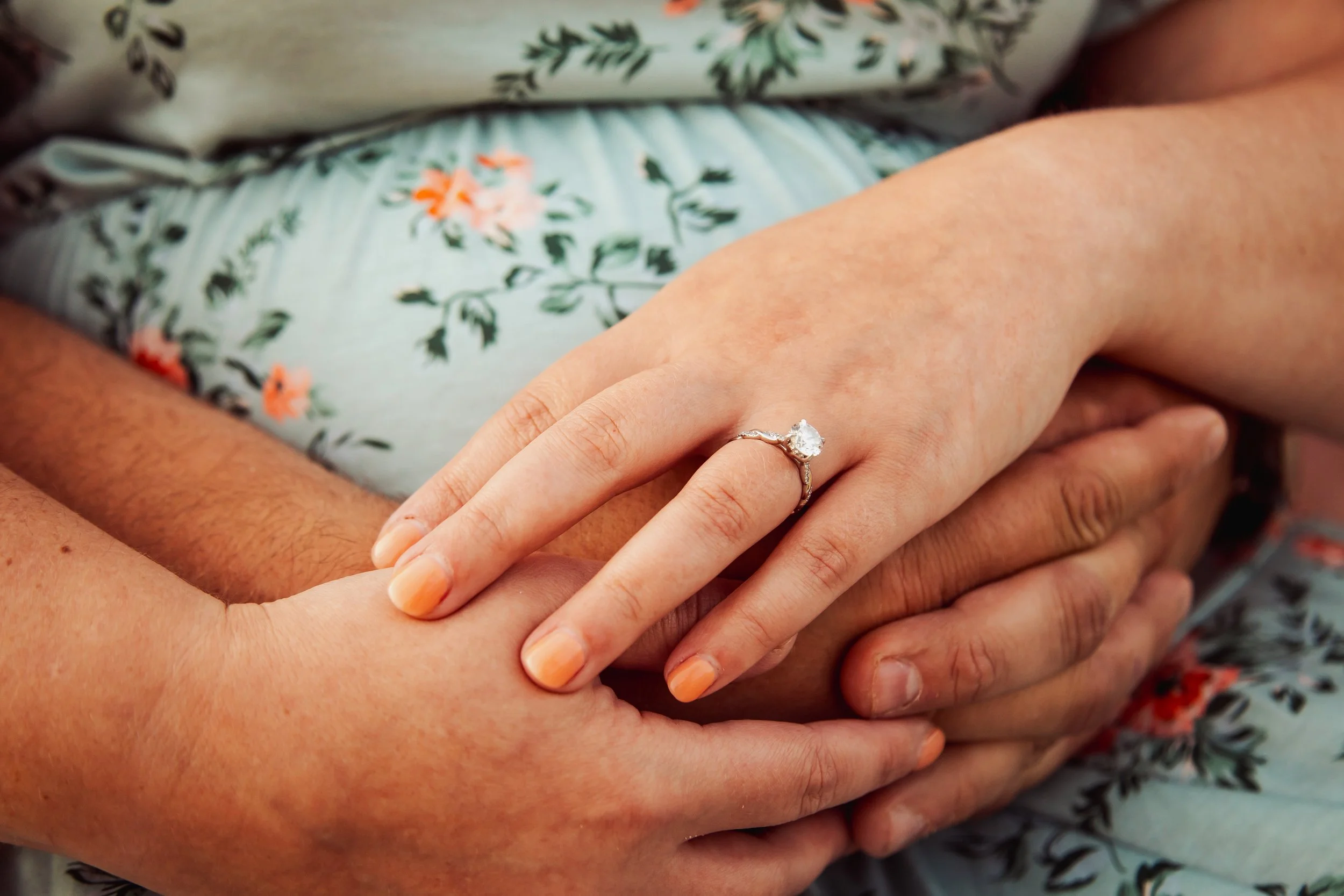 Close-up of two hands, one with an engagement ring featuring a large diamond, gently holding each other on the person's lap, who is wearing floral patterned clothing.