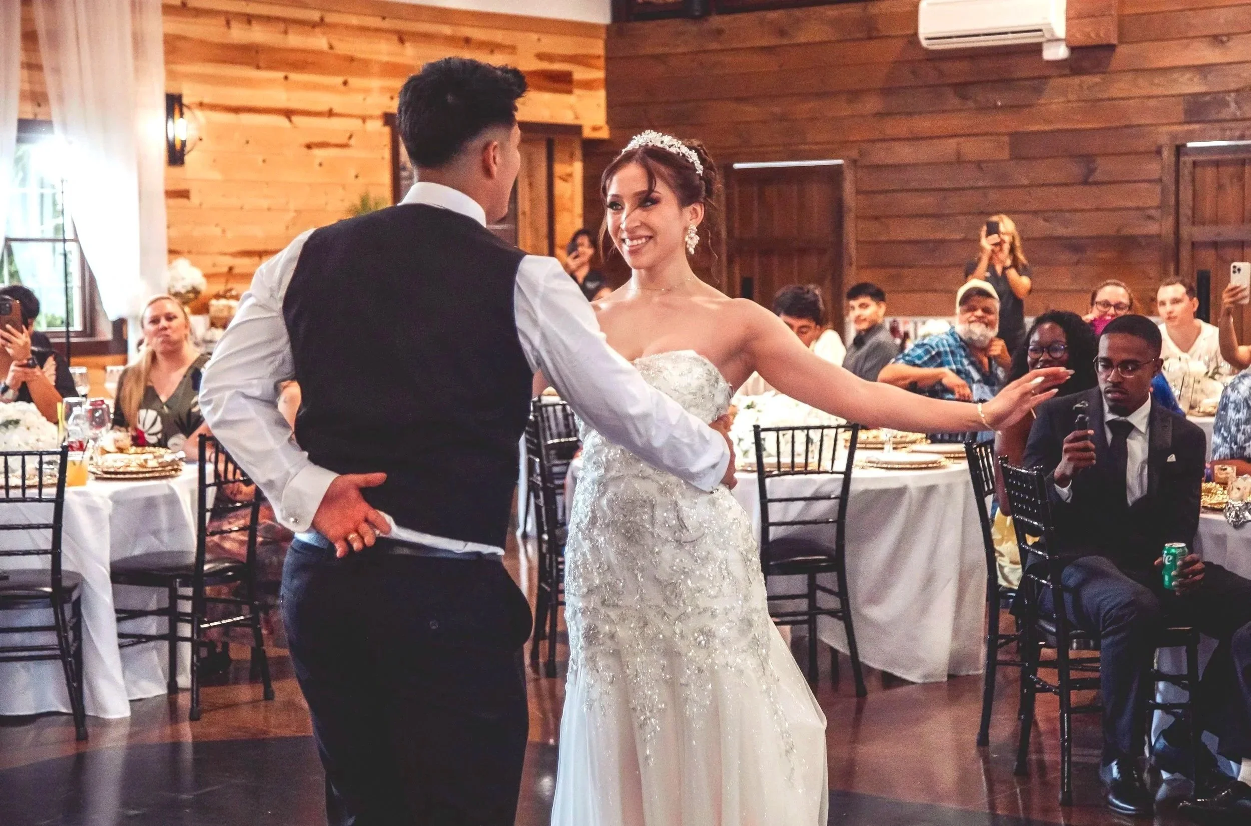 A bride and groom are dancing at their wedding reception in a rustic wooden hall. The bride is wearing a strapless white gown with intricate beadwork and a tiara. The groom is dressed in a black vest and white shirt. Guests are seated at round tables around them, some taking photos and videos, with a wood-paneled interior and large windows in the background.