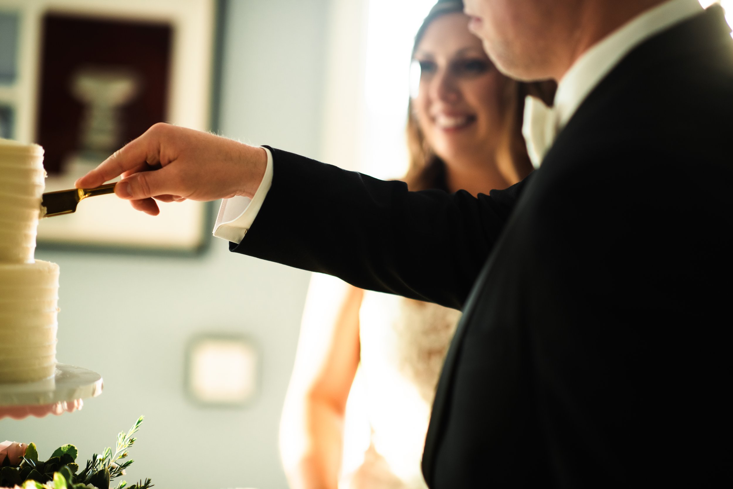 A man in a tuxedo and a woman in a dress cutting a wedding cake with a gold knife.