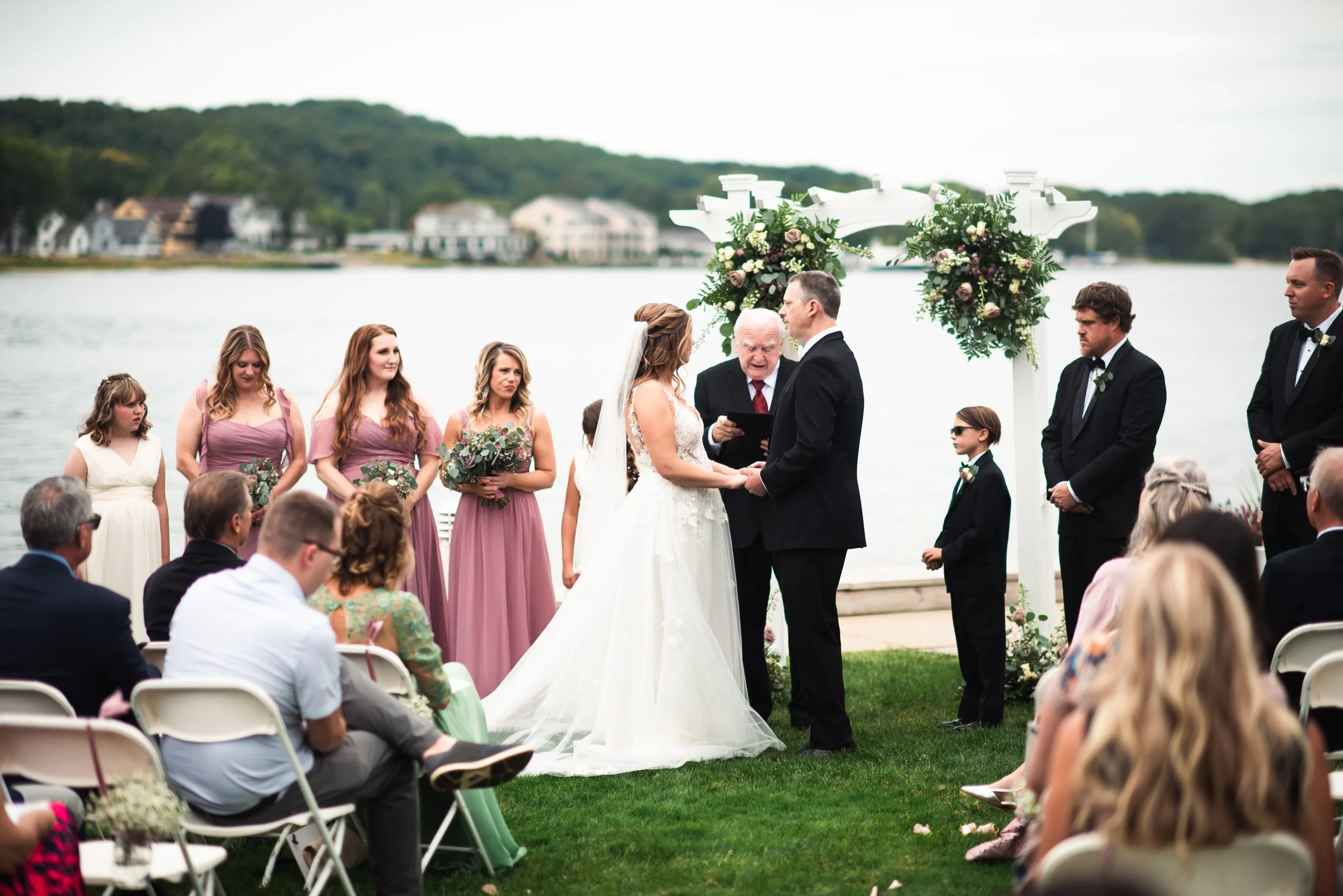 A wedding ceremony taking place outdoors by a lake with a white floral arch, where a bride and groom are exchanging vows. Bridesmaids in mauve dresses and groomsmen in black suits stand nearby, with guests seated in front watching the ceremony.