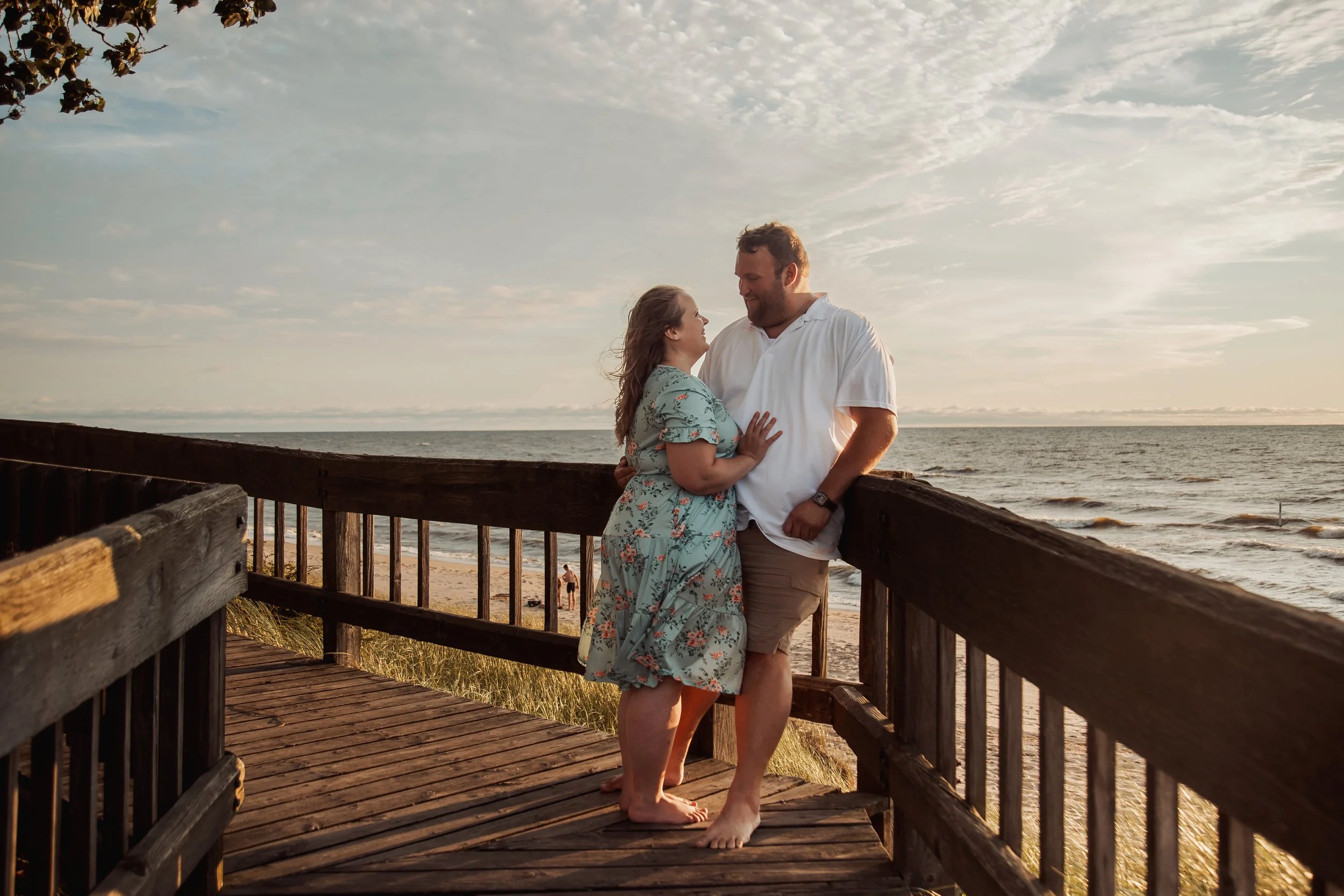 A couple standing on a wooden overlook by the beach, facing each other and smiling with the ocean in the background during sunset.