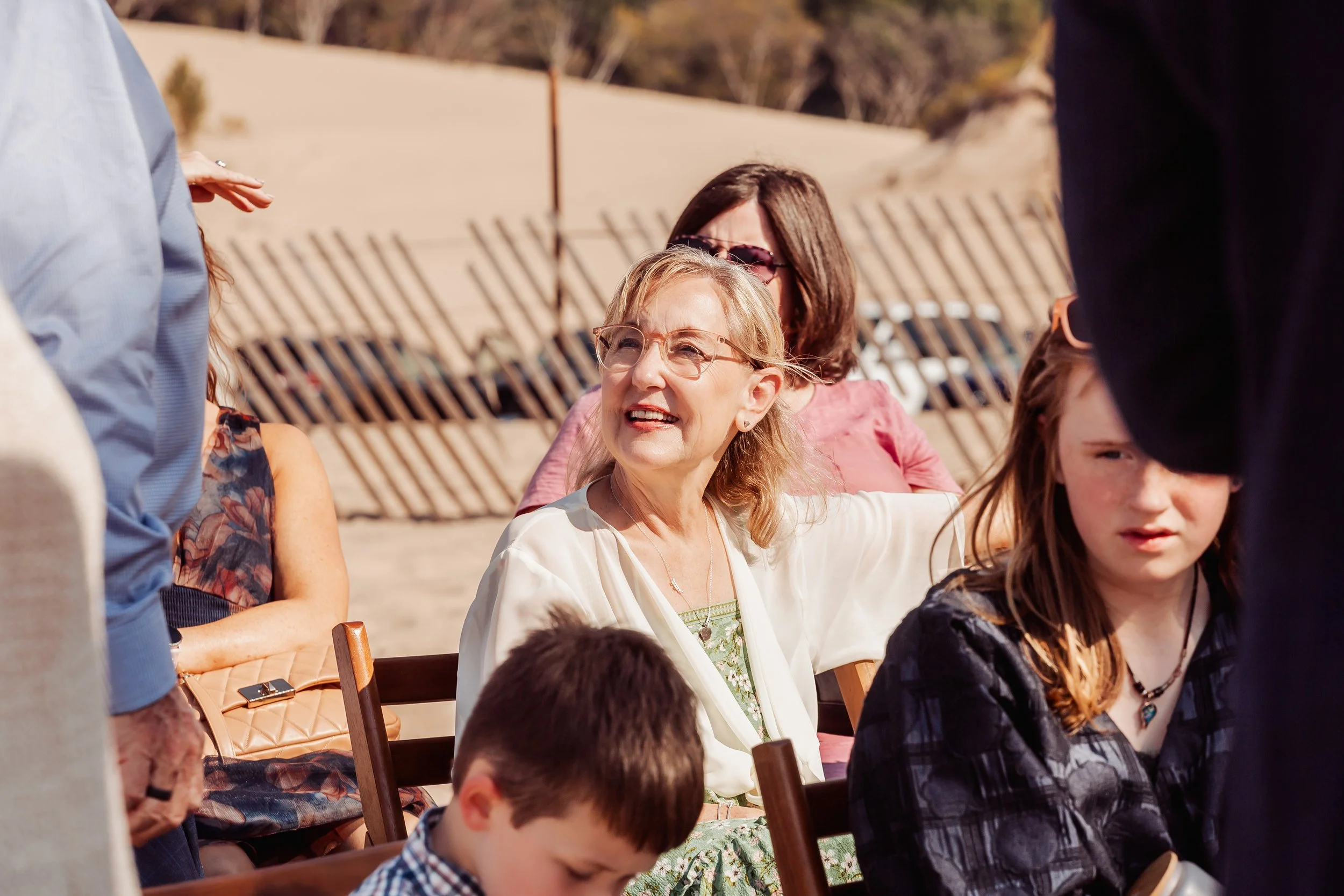 A group of people outdoors, including a woman with blonde hair, glasses, and earrings smiling and talking to someone off-camera, with children and other adults around.