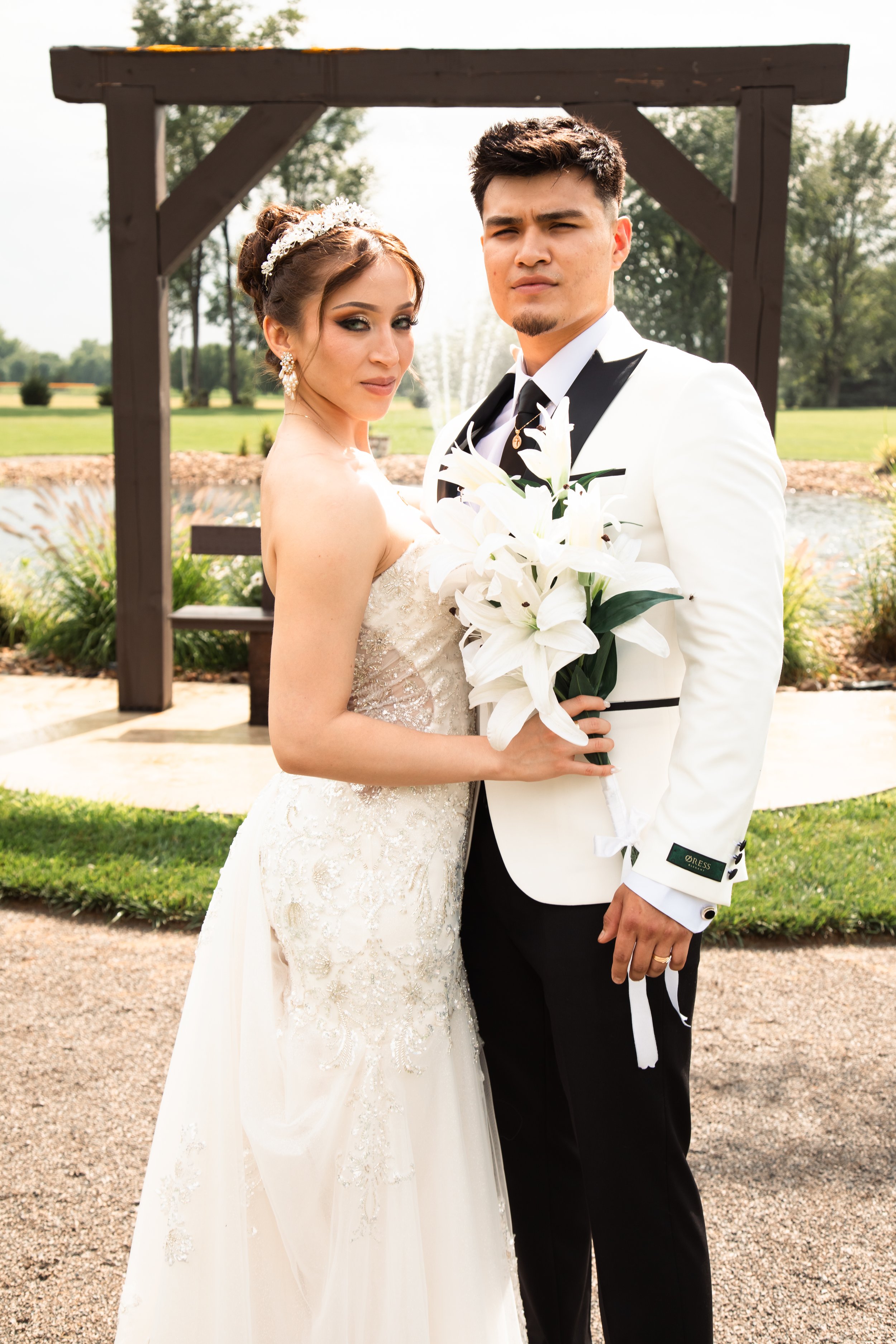 A bride and groom posing outdoors during their wedding, with the bride holding a bouquet of white lilies.