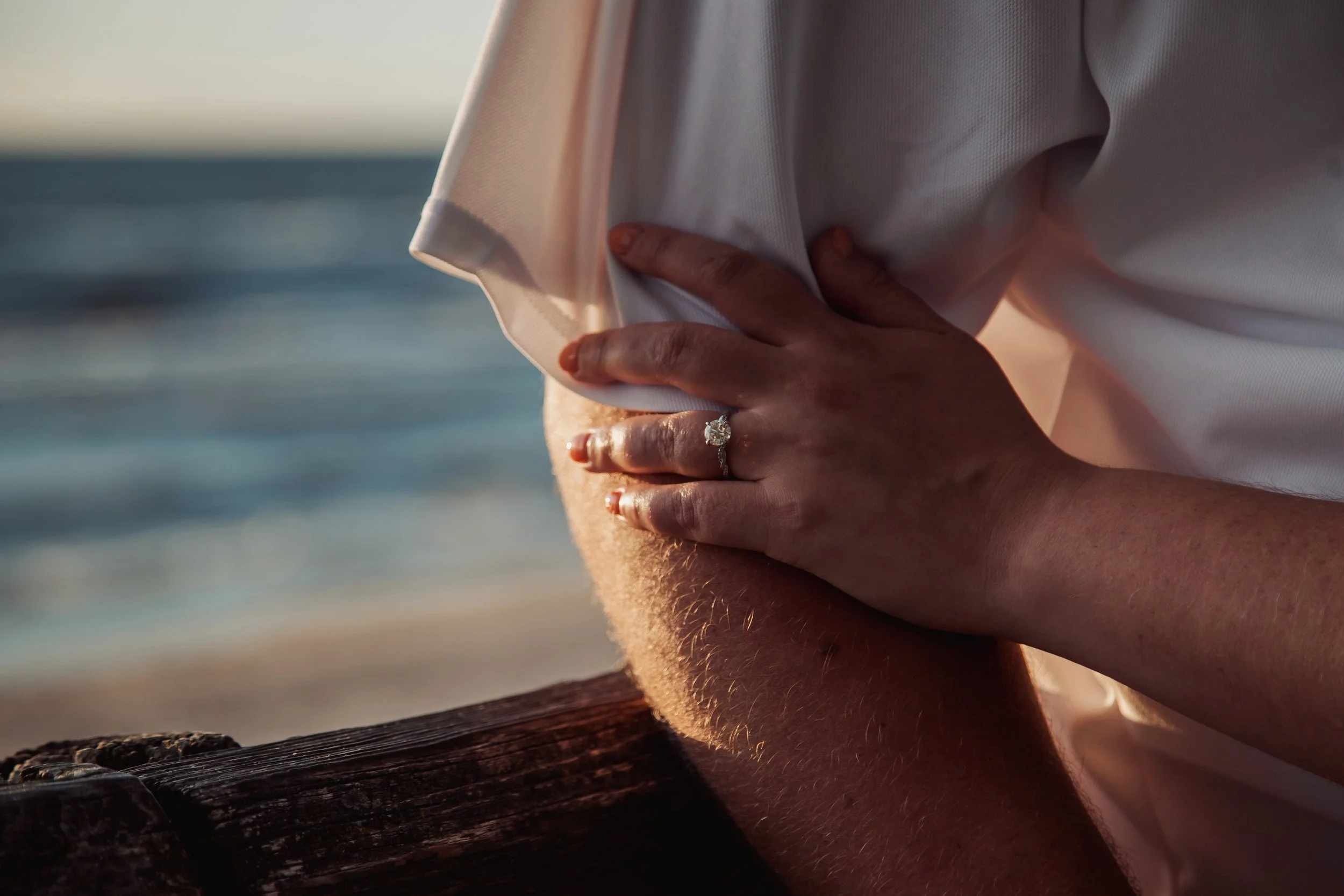 Close-up of a hand with a ring resting on someone's arm, with a blurred ocean background.