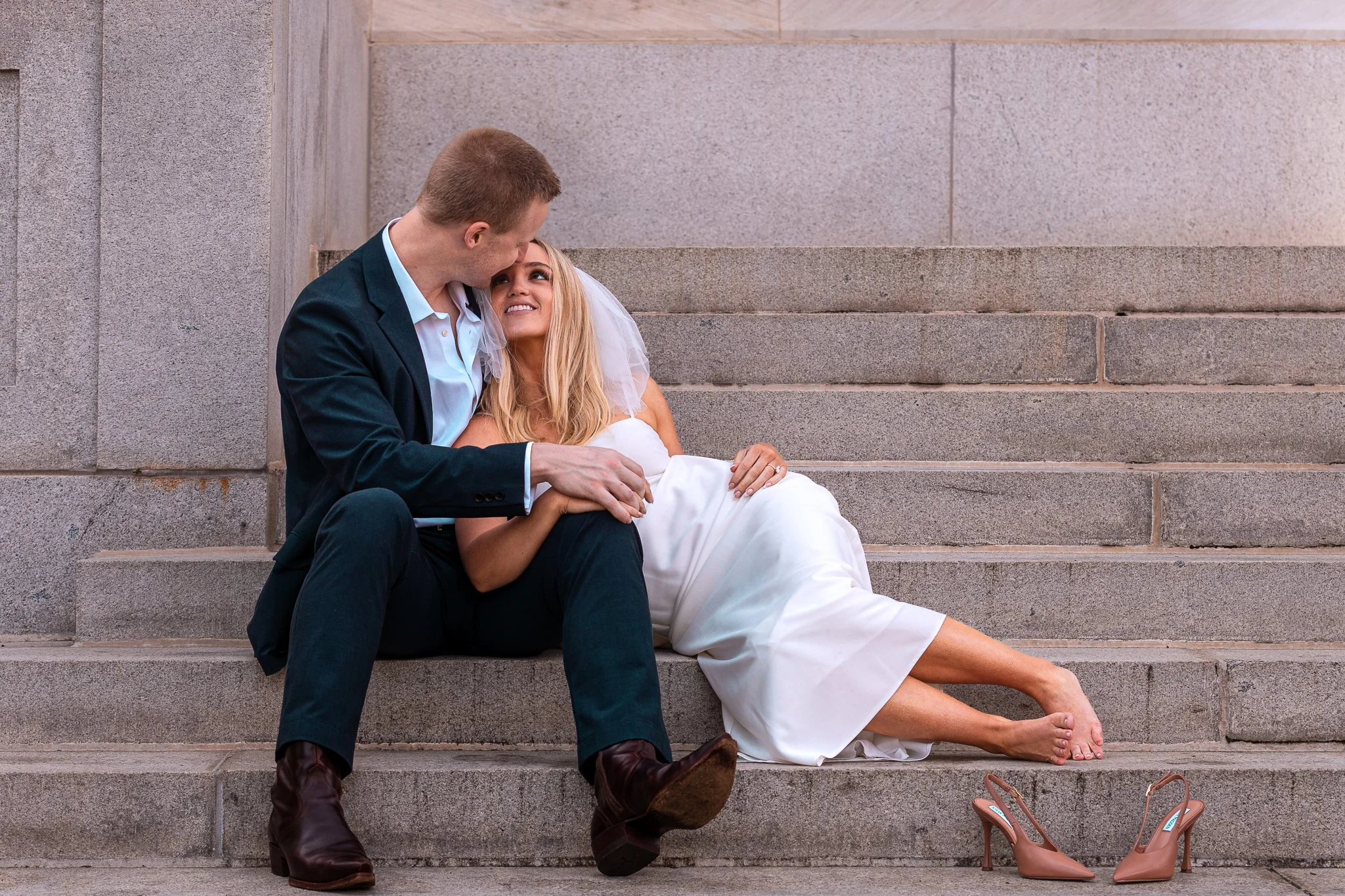 A man and woman, dressed for a wedding, sitting on steps, looking at each other, with the woman lying back on the steps and the man holding her. A pair of pink high-heeled shoes is on the steps beside them.