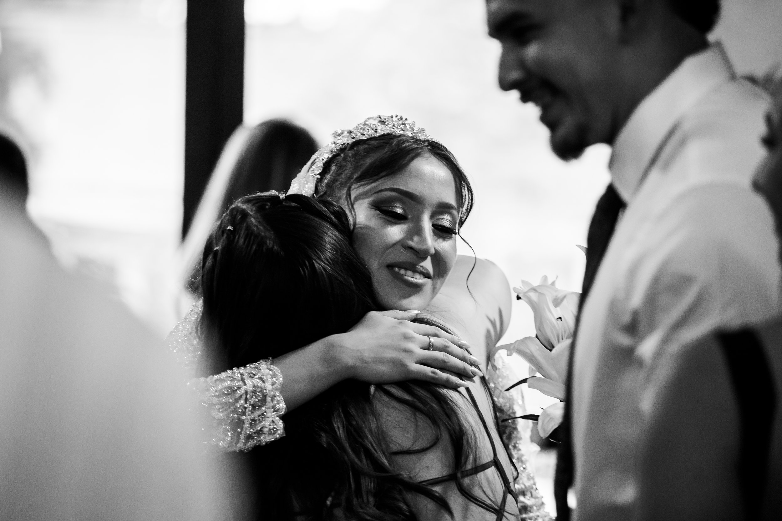 Black and white photo of a woman in a bridal gown hugging a young girl, with a man smiling nearby, at a wedding ceremony.