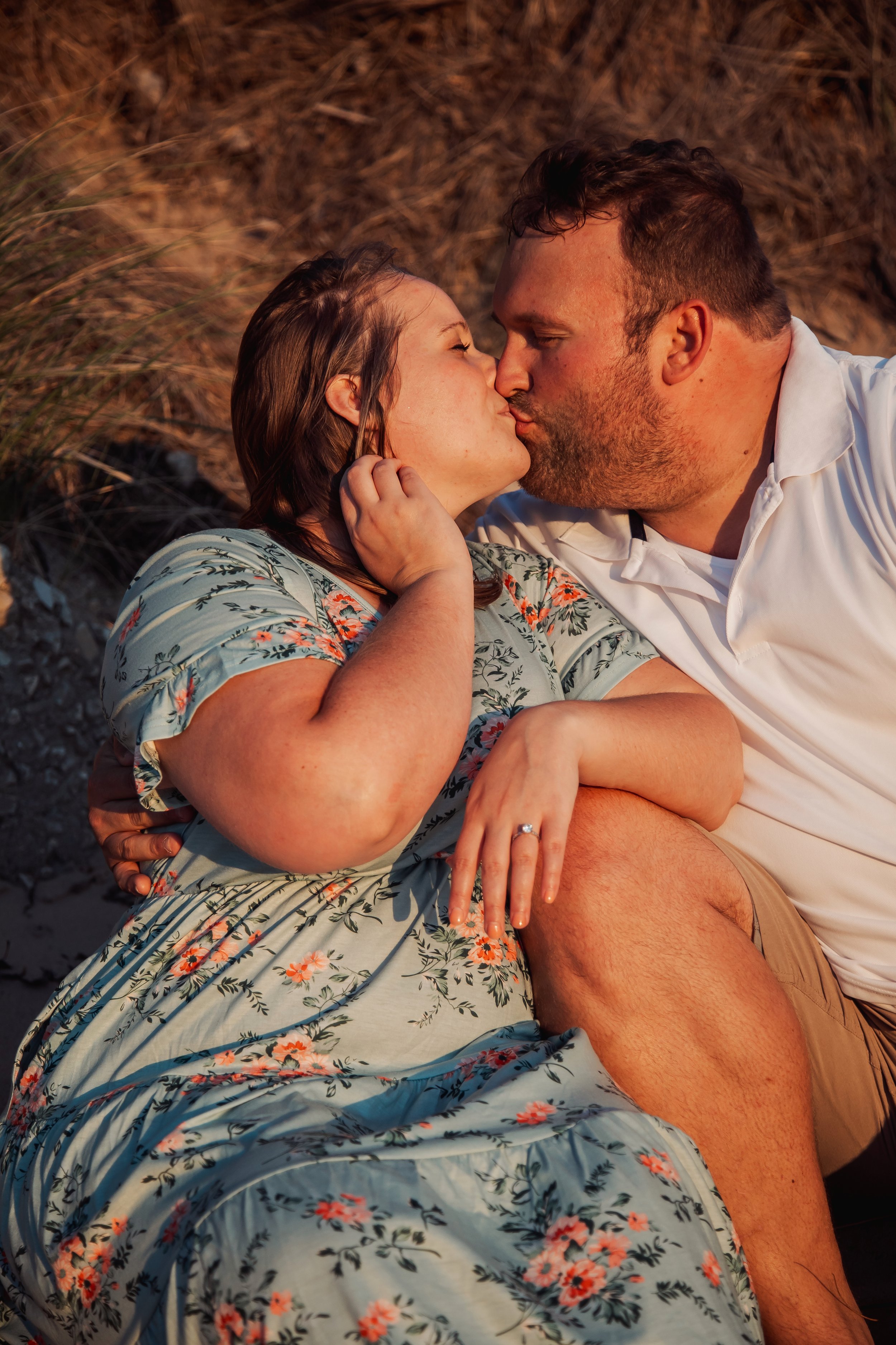 A couple is sharing a kiss on the beach, laying on sand near dry grass, during sunset.