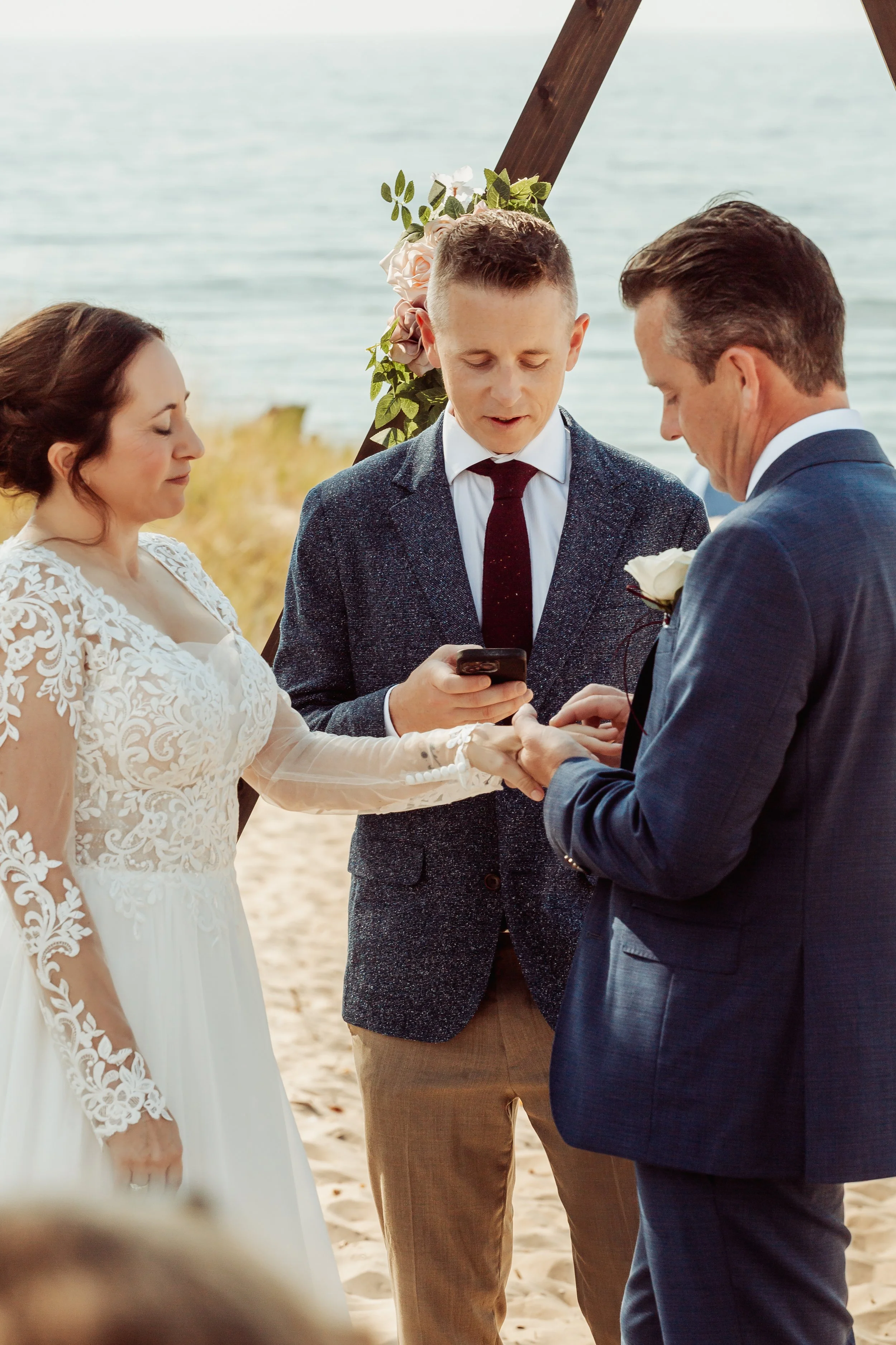 A wedding ceremony taking place outdoors on a beach, with a bride and groom holding hands and exchanging rings, overseen by an officiant.