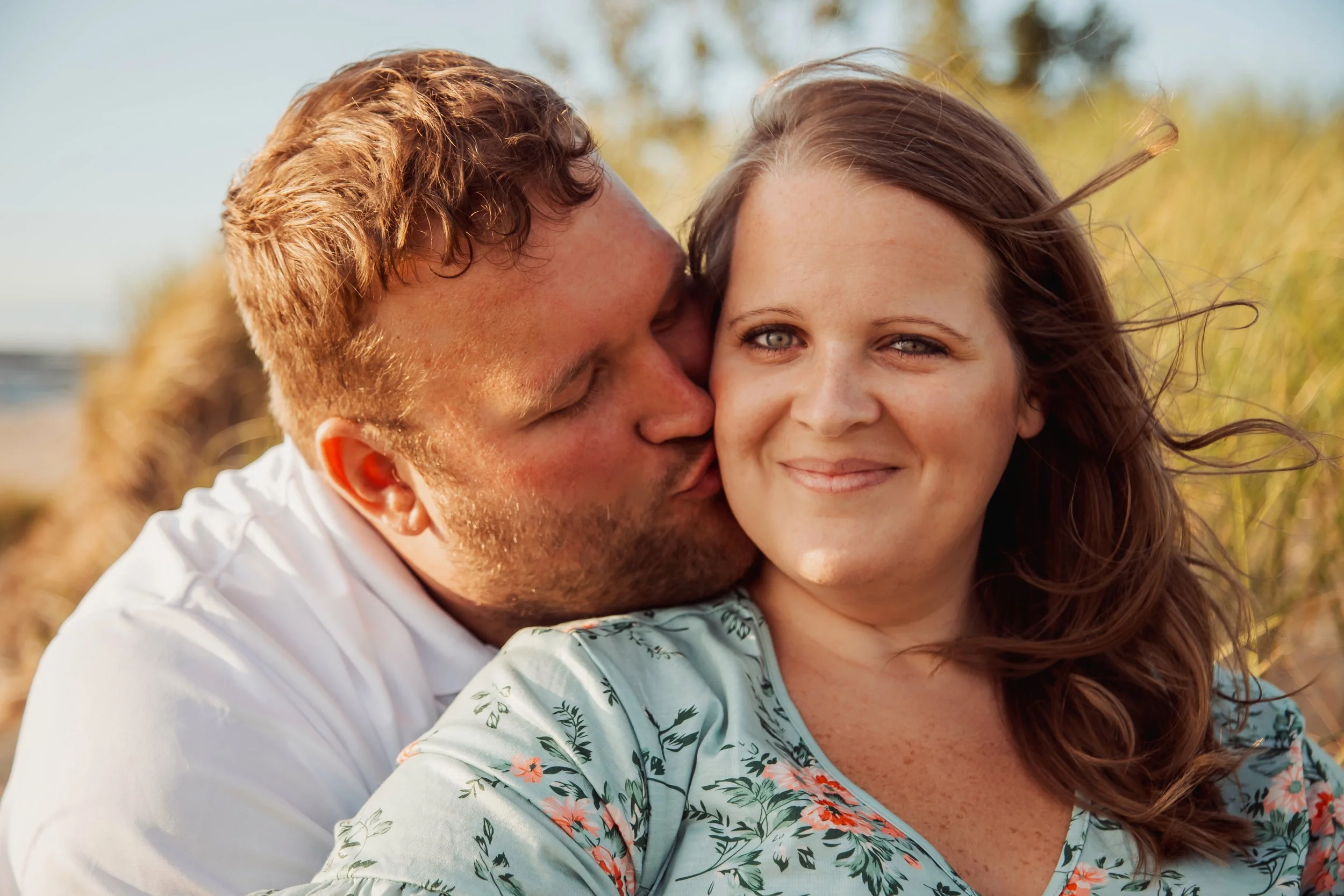 A man kisses a woman on the cheek at the beach with dunes and grass in the background.