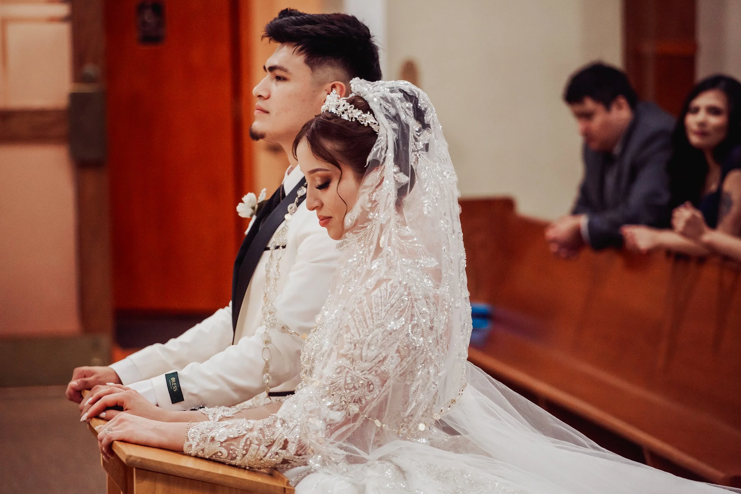 Bride and groom kneeling in prayer during their wedding ceremony, with guests in the background.