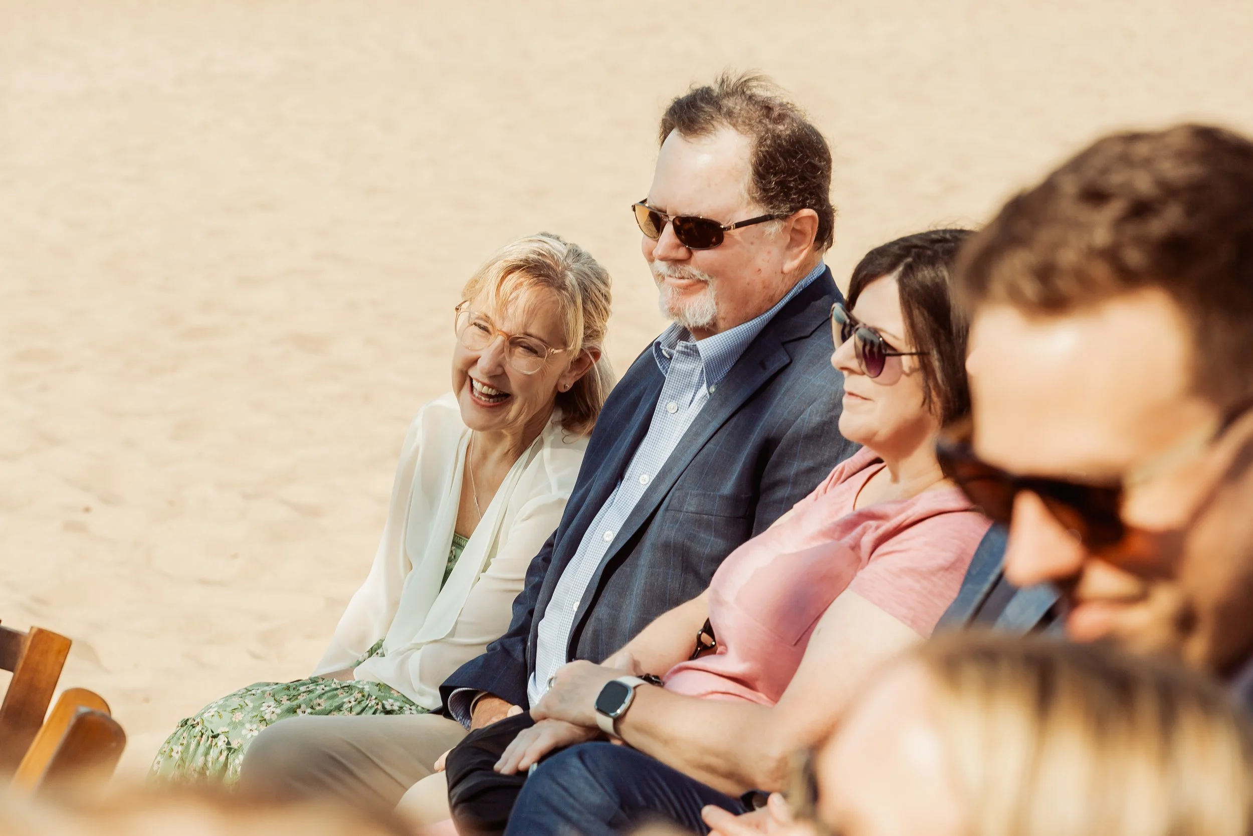 Group of four adults sitting on benches at a beach, smiling and enjoying the sunny weather.