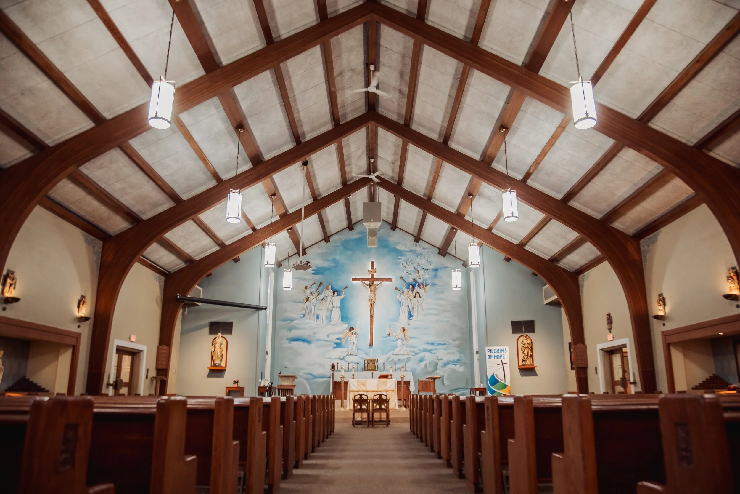 Interior of a church with wooden pews, a painted mural of angels and clouds behind the altar, and hanging pendant lights.