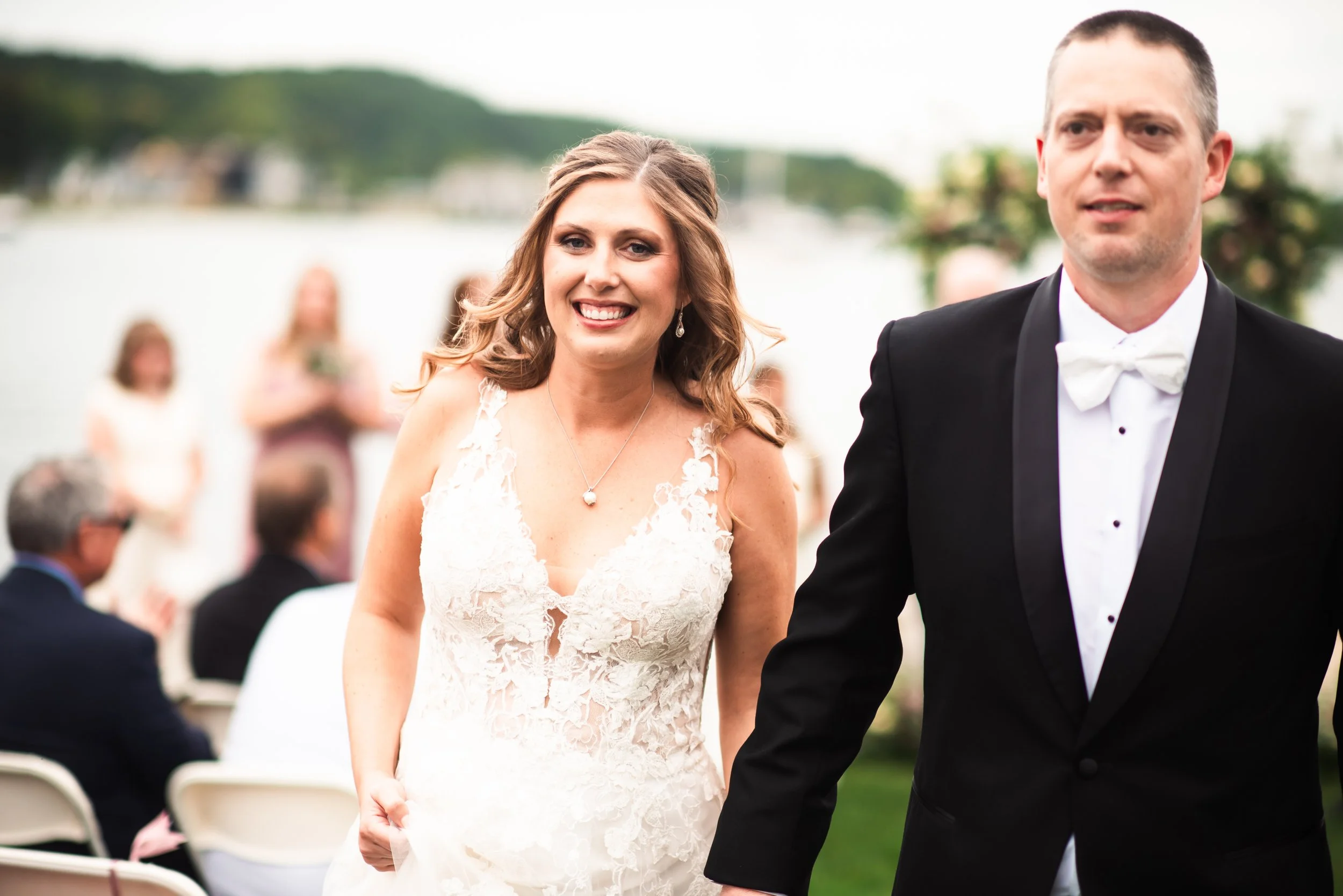 A smiling bride in a white lace wedding dress holding hands with a groom in a black tuxedo during an outdoor wedding ceremony by the water.