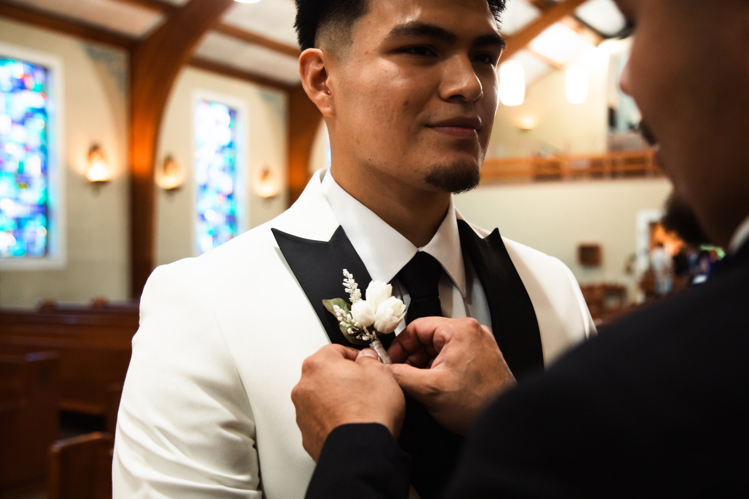 A man in a white tuxedo with a black bow tie is being helped to pin a white boutonniere onto his lapel by another person. The setting is inside a church with stained glass windows.