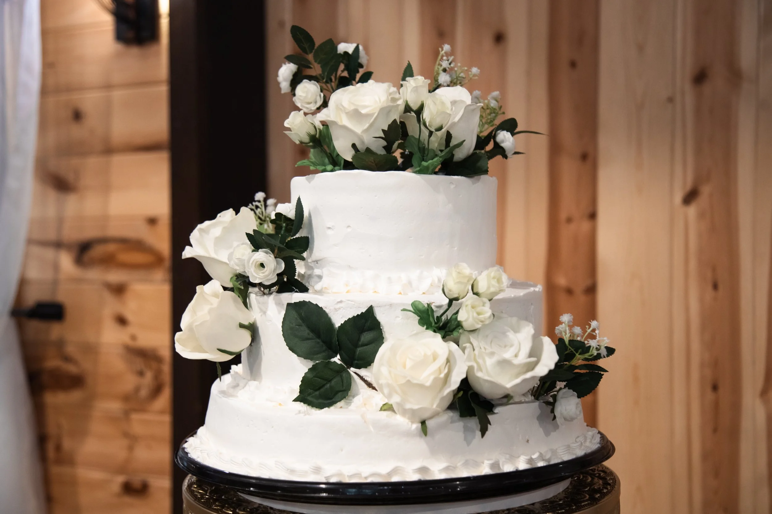 A three-tier white wedding cake decorated with white roses, greenery, and some small white flowers, set on a black cake stand against a wooden wall background.